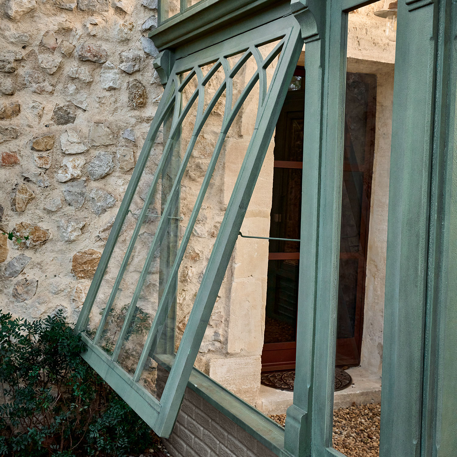 Grünes Metallfenster mit dekorativem Muster vor Steinmauer und Pflanzen im Hintergrund