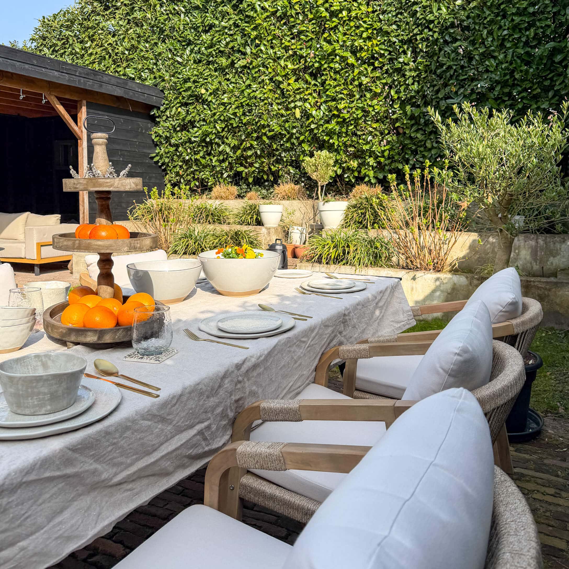 Gartentisch mit weißem Leinentuch, Keramikgeschirr und hölzernem Obstständer bei Sonnenschein