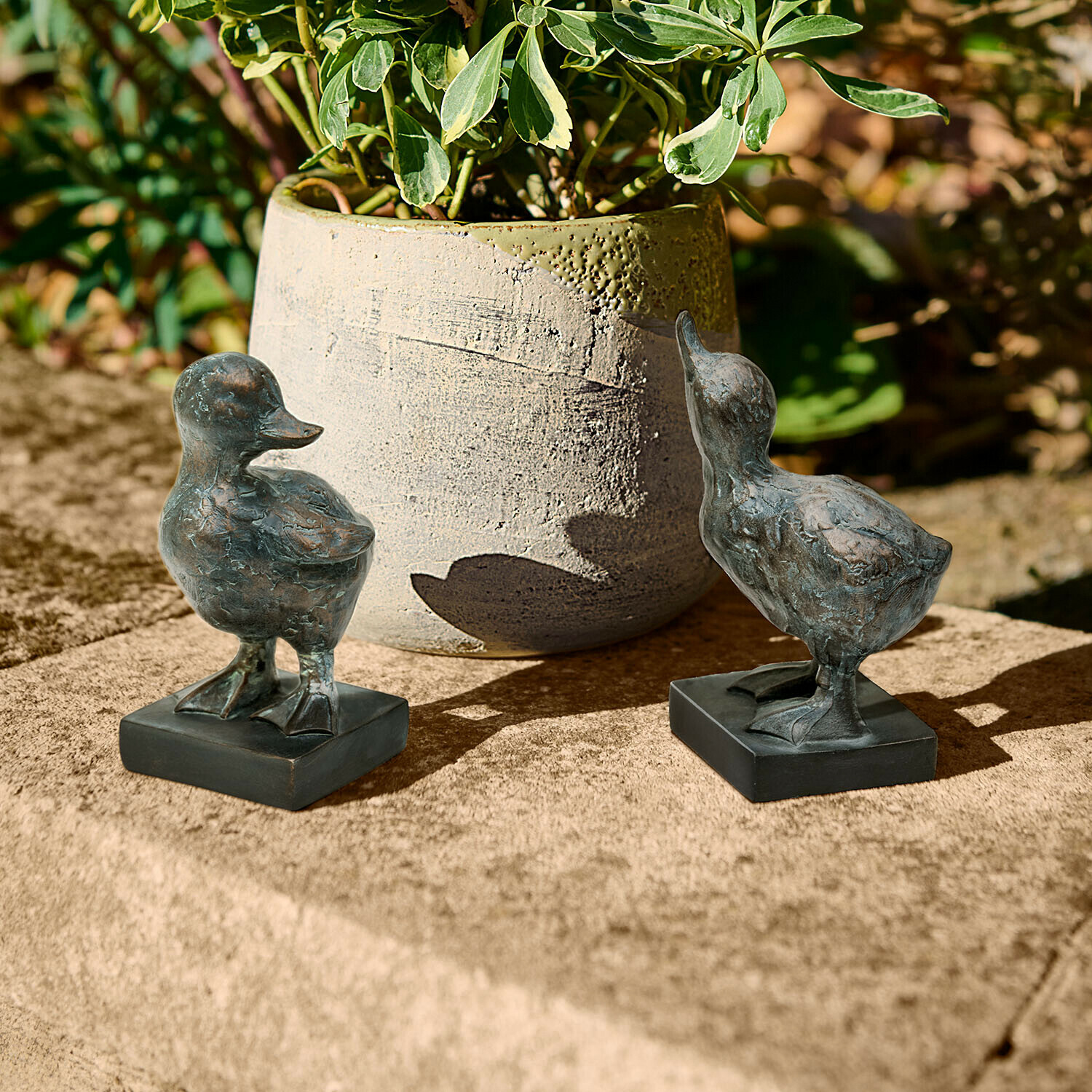 Deko-Gänseküken 2er Set Merrowden Zwei dunkle Metall-Entenfiguren auf Steinplatte neben einer Pflanzvase im Garten