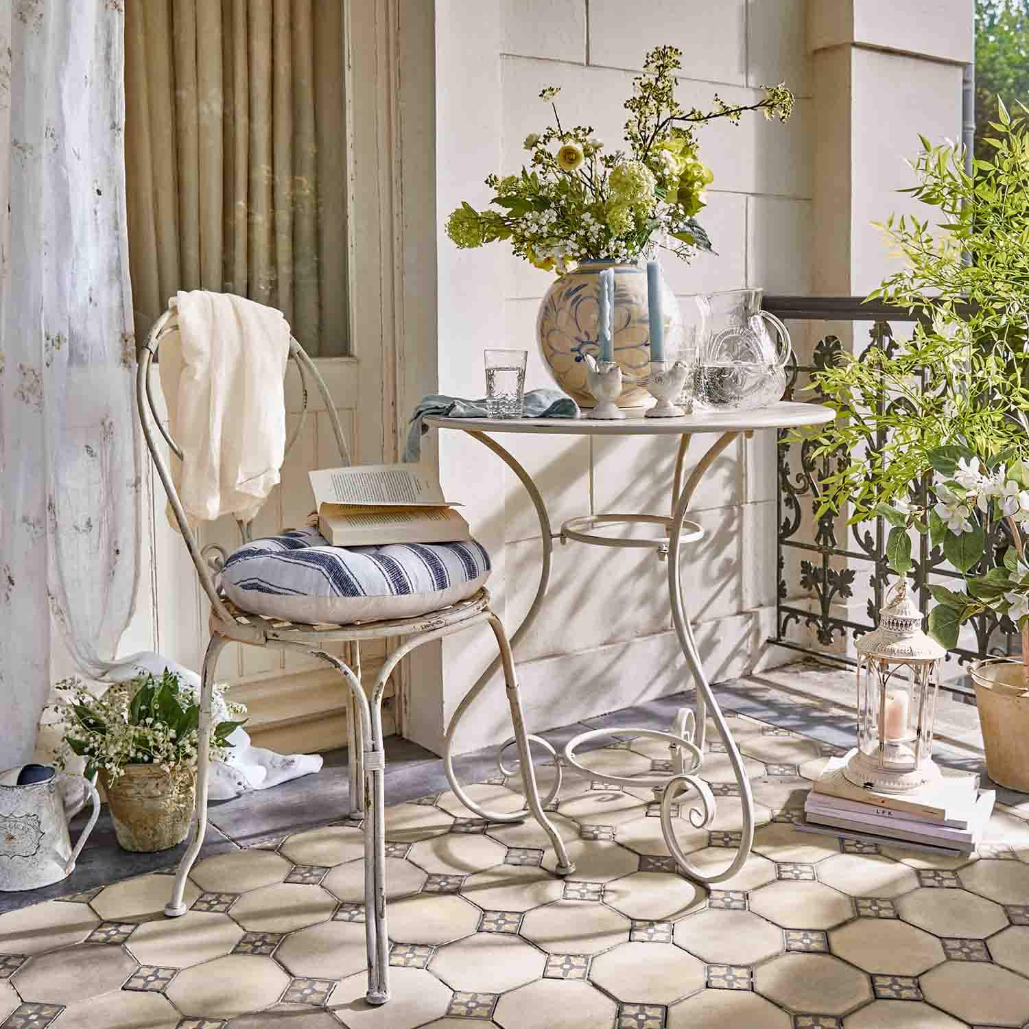 Weißer Metalltisch und Stuhl mit gestreiftem Kissen auf Terrasse mit Blumen und Laterne