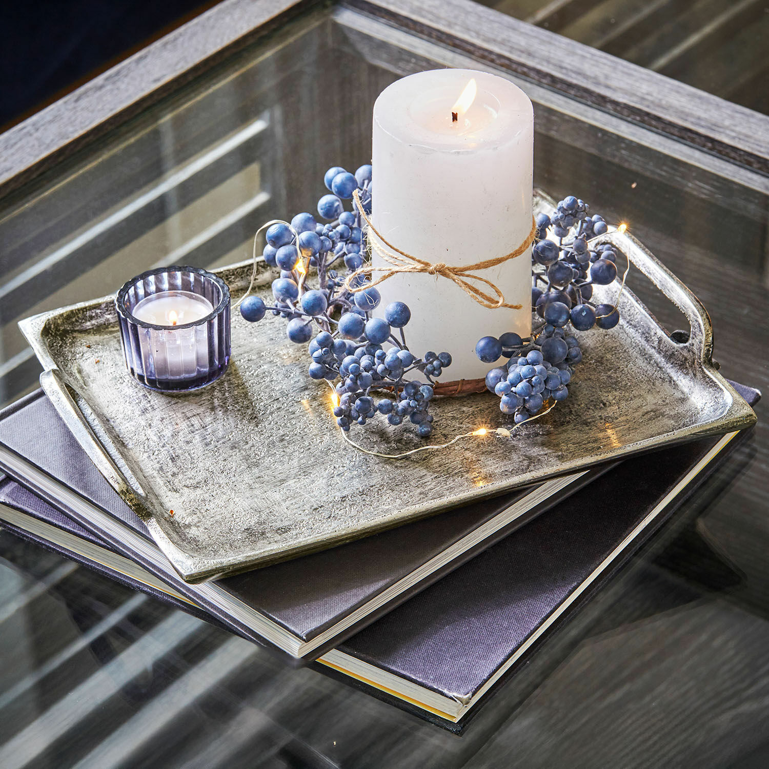 Weiße Kerze mit Naturkordel und blauen Beeren auf Metalltablett auf Glastisch dekoriert