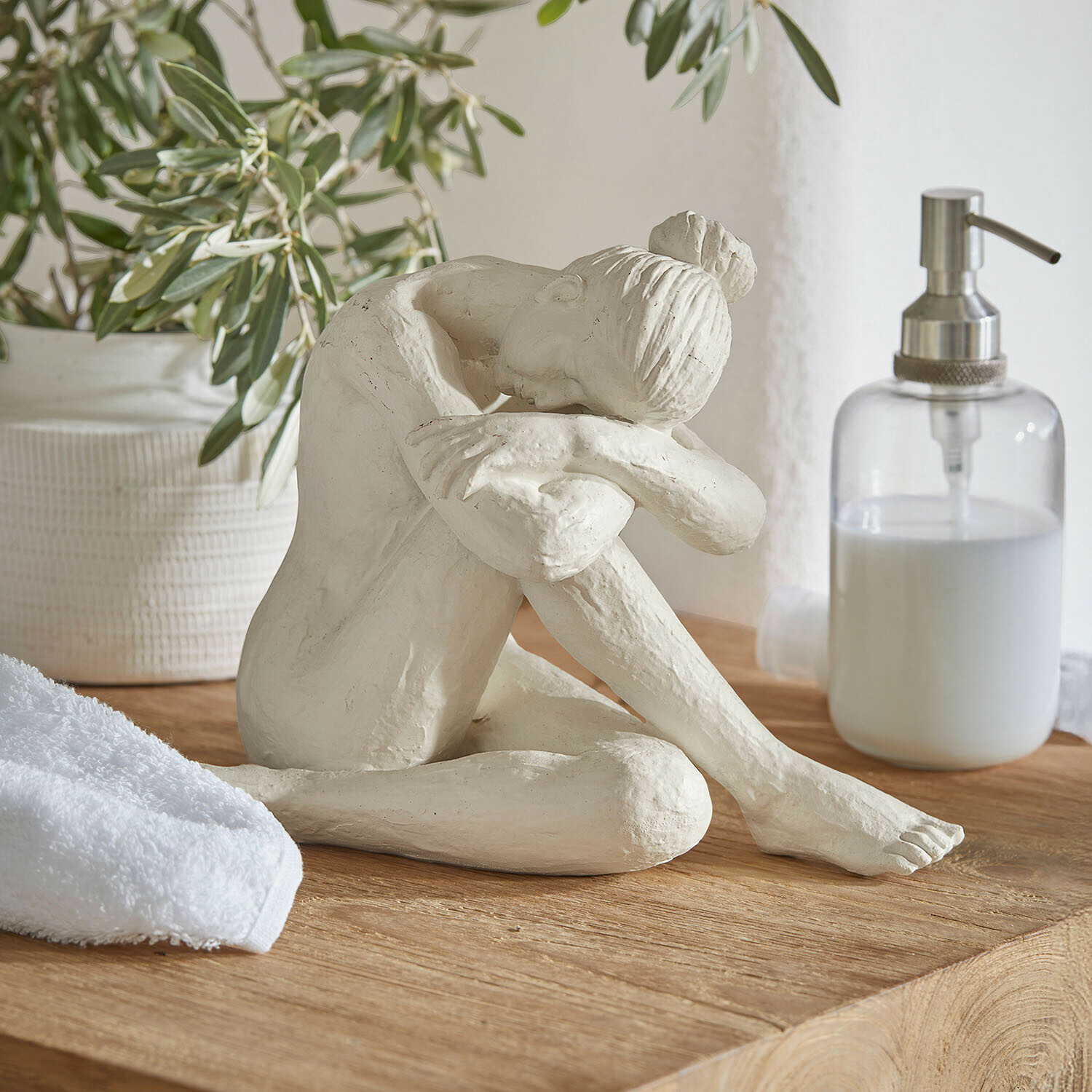 Weiße Skulptur einer sitzenden Frau mit Kopf auf angewinkelten Knien auf Holzplatte im Badezimmer