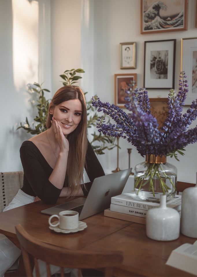 Frau an Holztisch mit Laptop und Glasvase mit violetten Blumen in hellem Raum