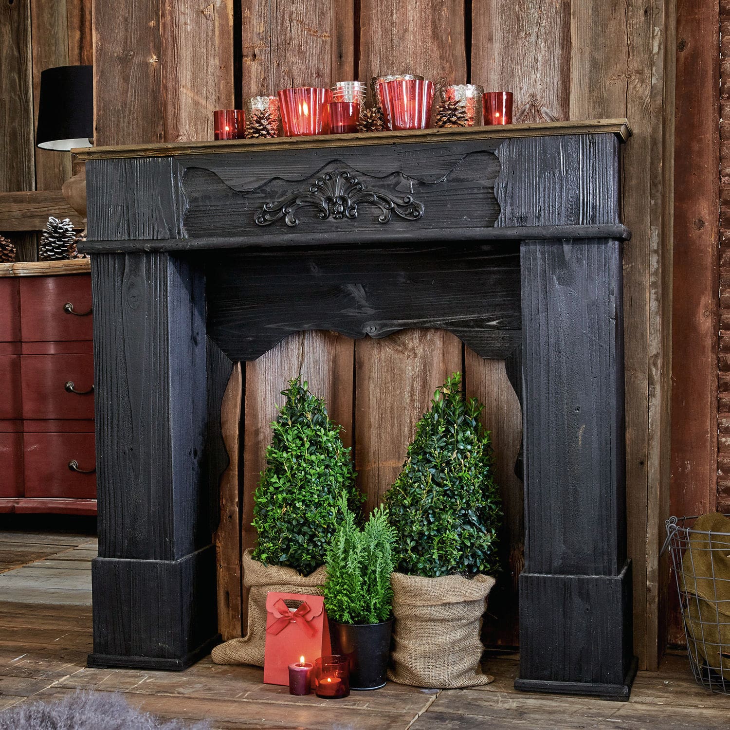 Kaminkonsole Margaux Schwarzer Holz-Dekokamin mit grünen Pflanzen und roten Kerzen in rustikalem Raum
