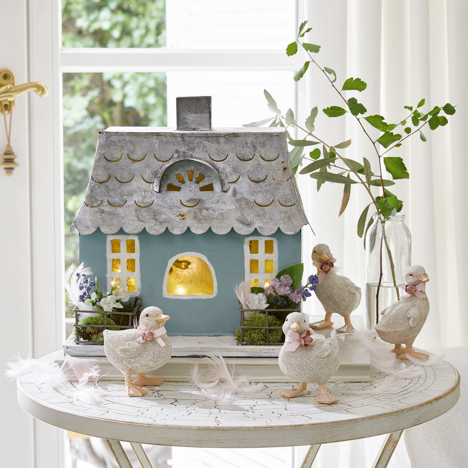 Dekoratives blaues Metallhaus mit vier weißen Entchen und Blumen auf rundem weißen Tisch