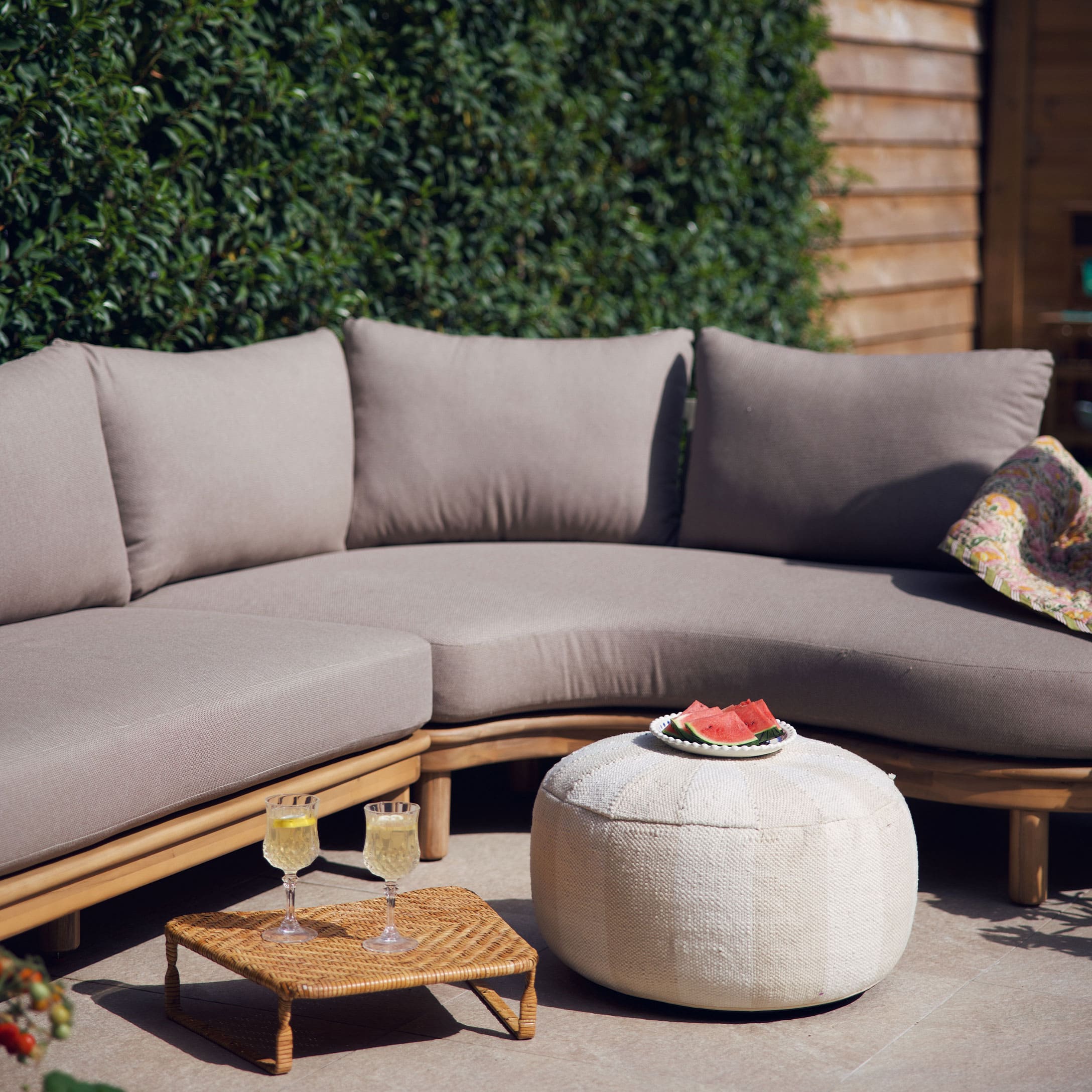 Beige Sofa mit Kissen auf Holzrahmen und weißer Pouf auf Terrasse mit sommerlicher Dekoration