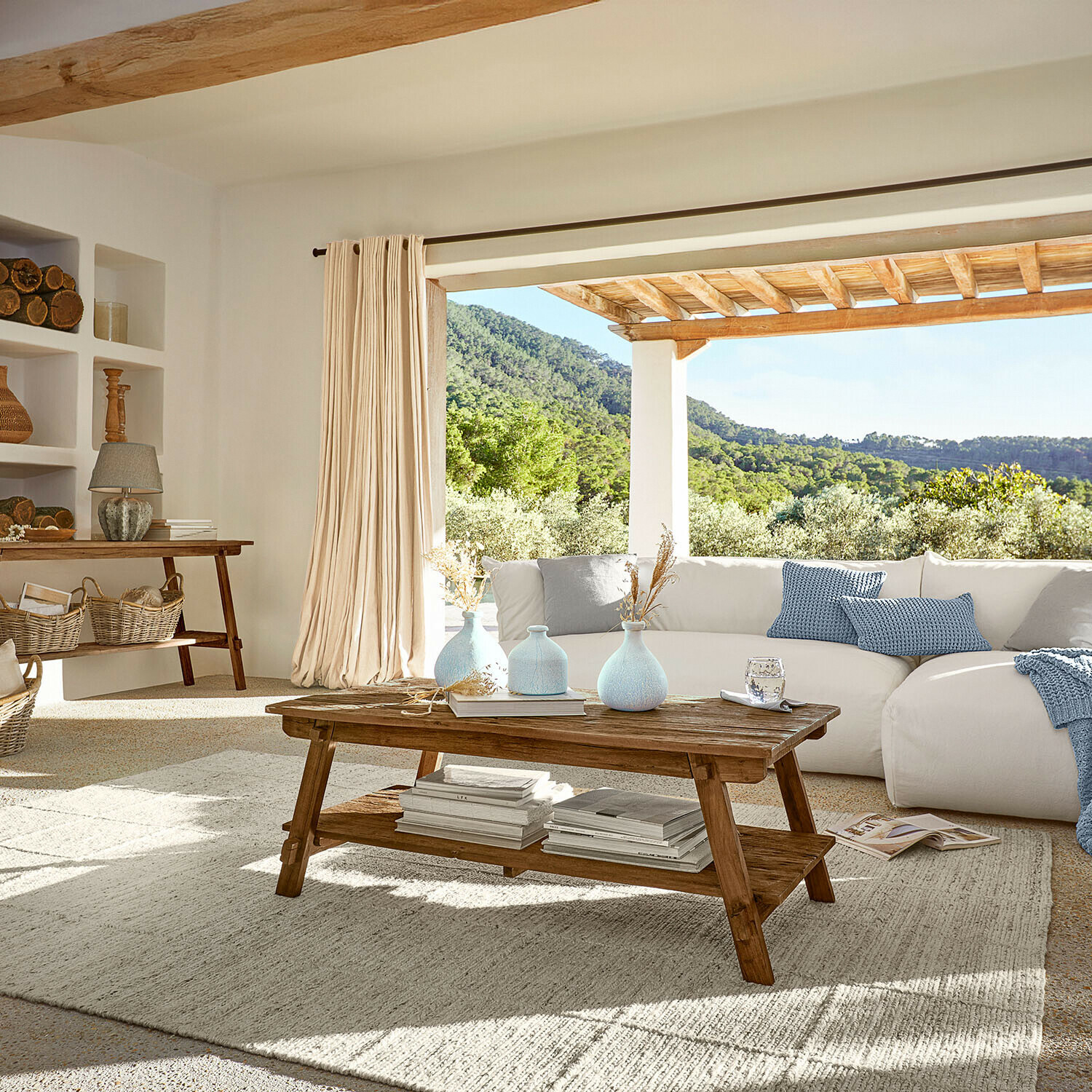 Wohnzimmer mit Holzcouchtisch und weißem Sofa mit blauen Kissen, natürlicher Lichteinfall und grüner Ausblick
