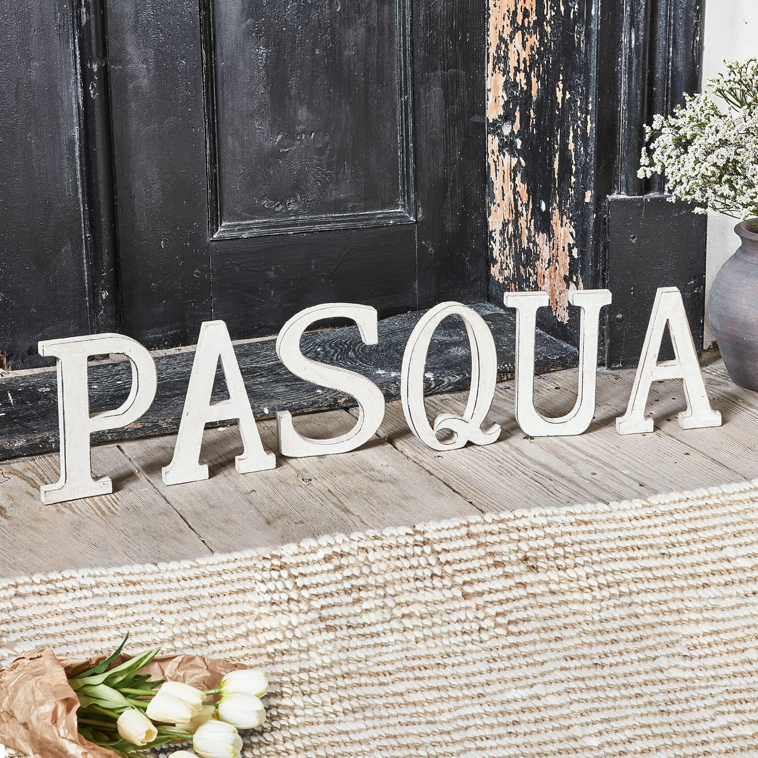 Deko-Buchstaben PASQUA Weiße Holzbuchstaben als Dekoration mit dem Schriftzug Pasqua auf Holzboden vor schwarzer Tür