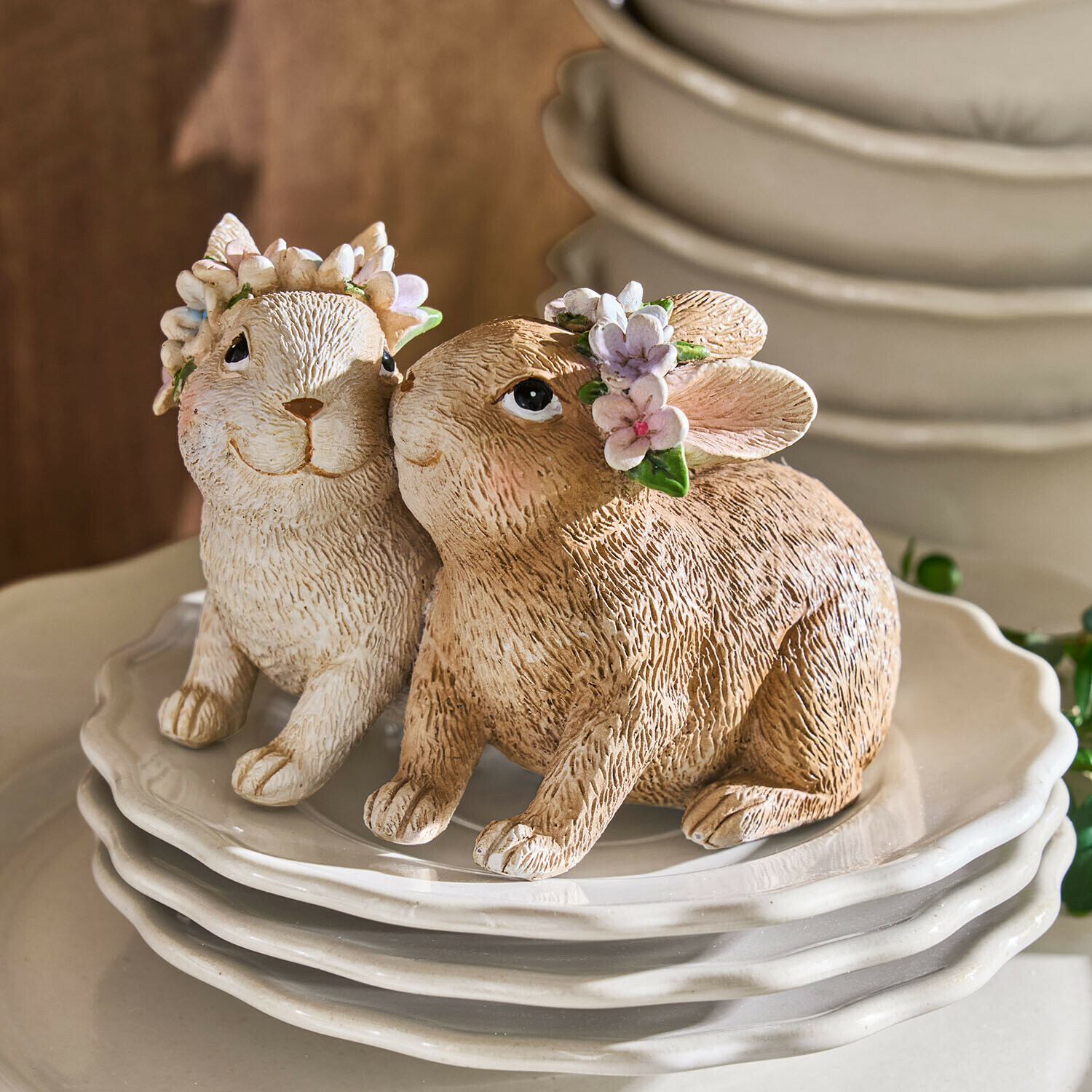 Zwei Keramik-Hasenfiguren in Beige und Weiß mit Blumenkränzen auf weißen Tellern