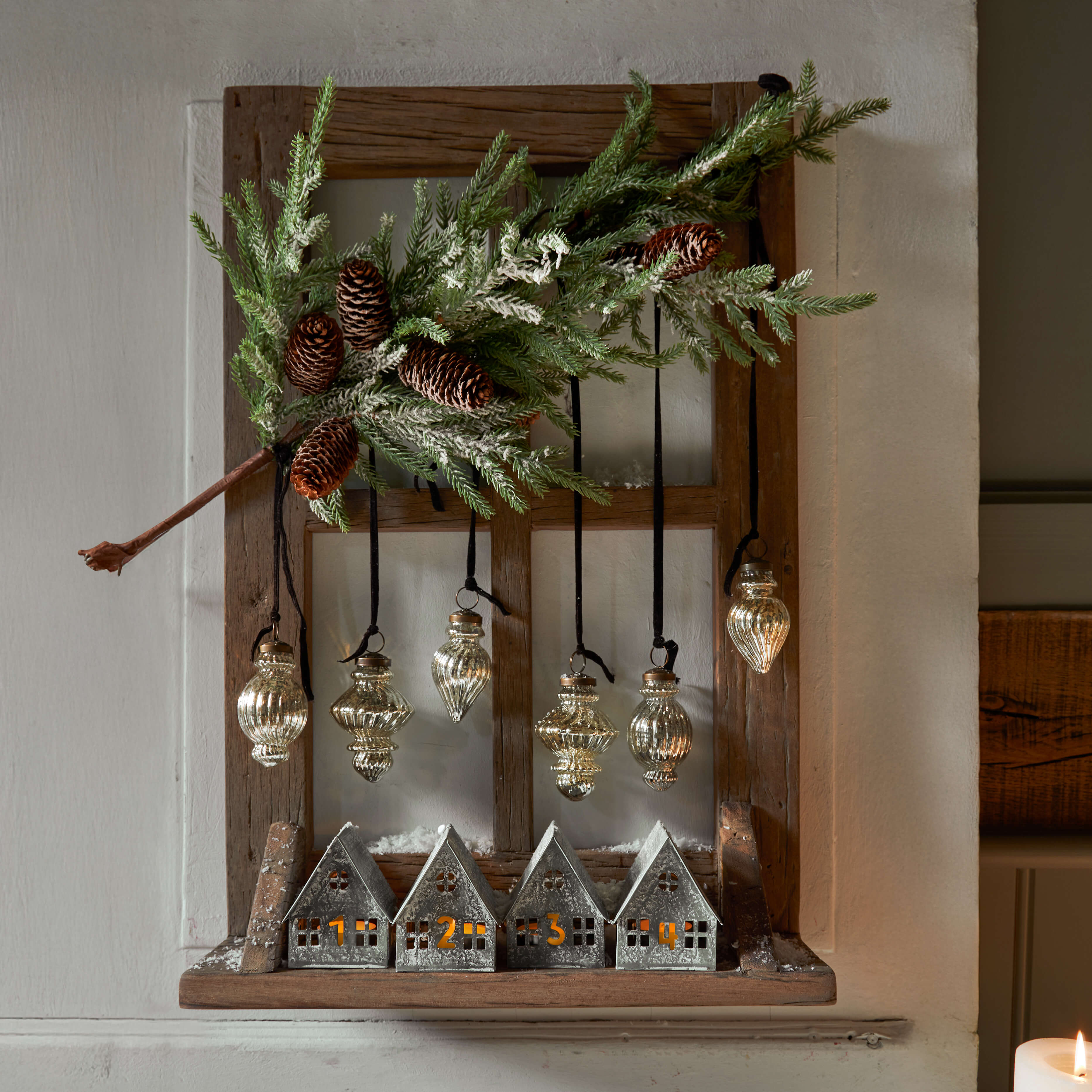 Holzrahmen mit nummerierten Metallhäusern und hängenden Glasornamenten, Weihnachtsdeko mit Tannenzapfen und grünen Zweigen