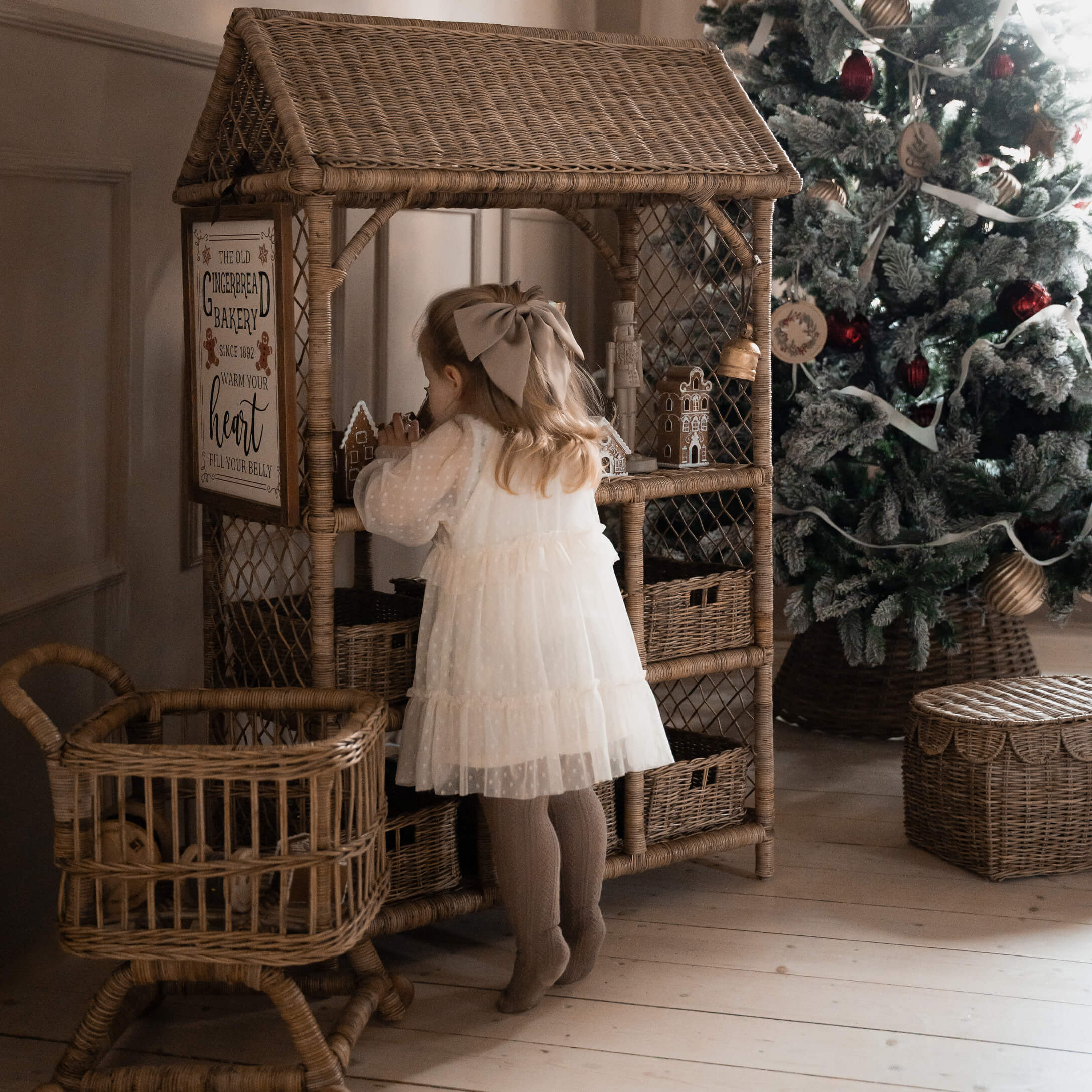 Kind in weißem Kleid vor einem Rattanschrank mit Lebkuchenhäusern neben geschmücktem Weihnachtsbaum