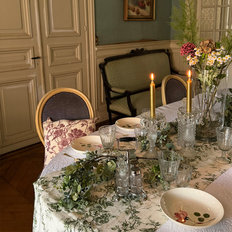 Eleganter Tisch mit weißem, grün gemustertem Tischtuch, brennenden Kerzen und Blumenarrangement im klassischen Raum