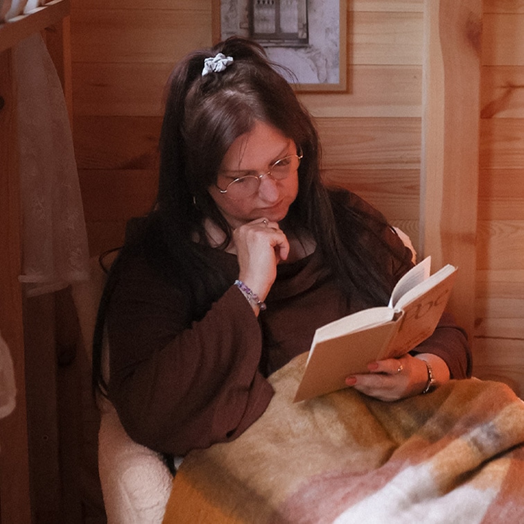Frau liest Buch unter weicher, mehrfarbiger Decke in gemütlichem Holzinterieur, entspannter Moment