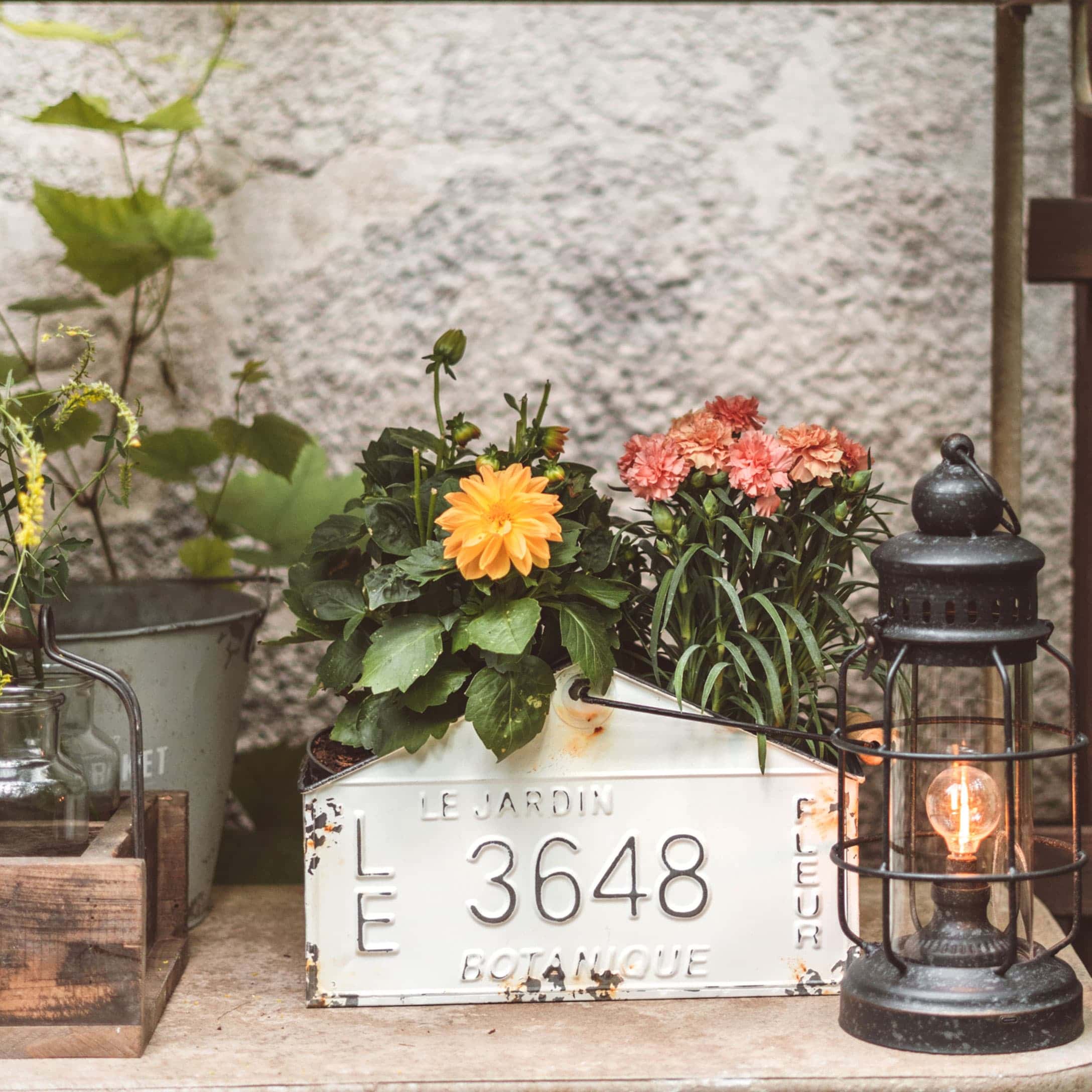 Weißer Metallbehälter mit Nummer 3648, bunten Blumen und schwarzer Laterne auf Gartentisch