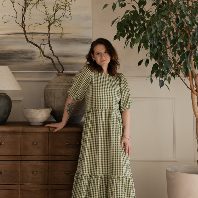 Frau in grün kariertem Kleid lehnt an Holzkommode in gemütlichem Raum mit Pflanzen