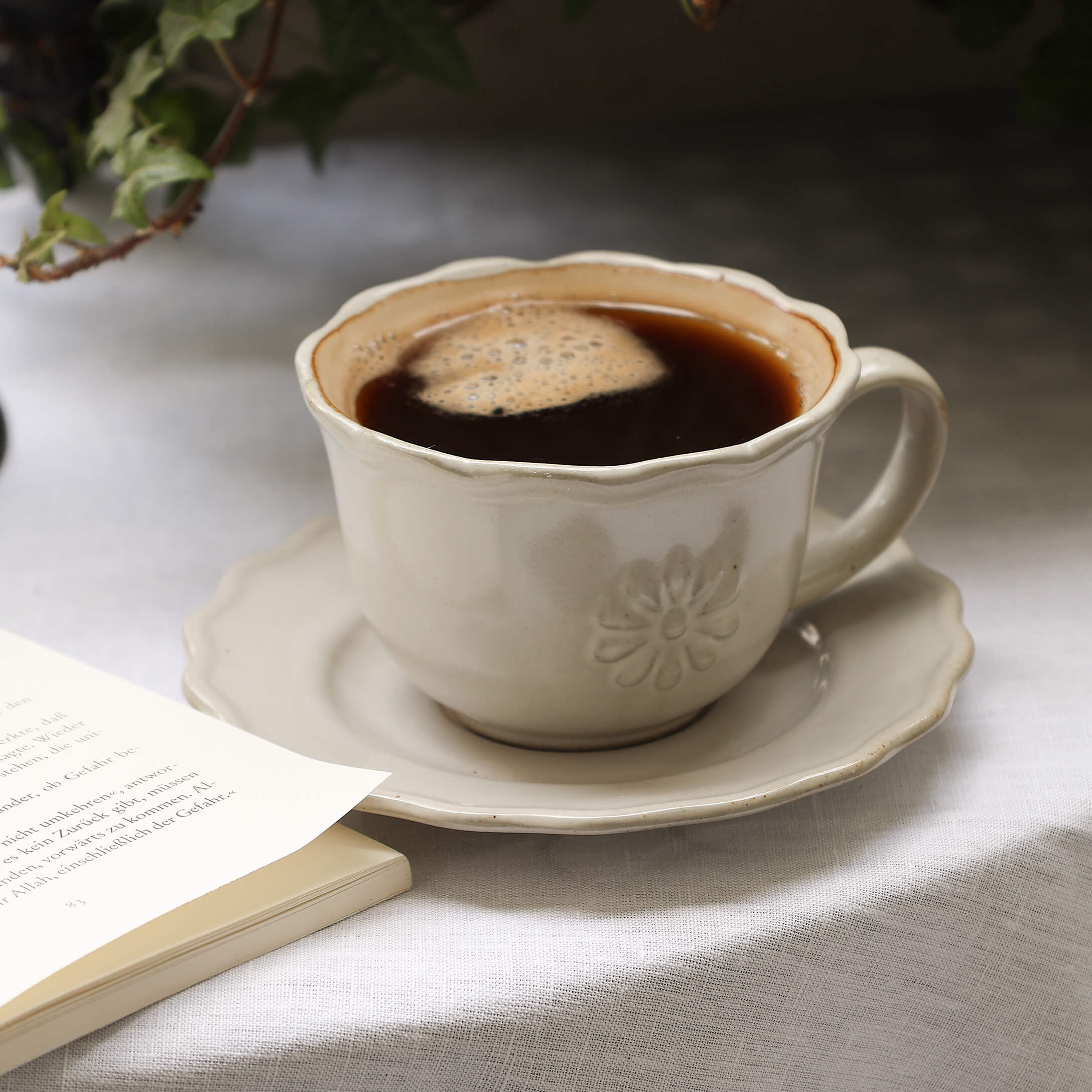 Beige Keramiktasse mit Kaffee auf Untertasse auf weißem Tischtuch neben offenem Buch
