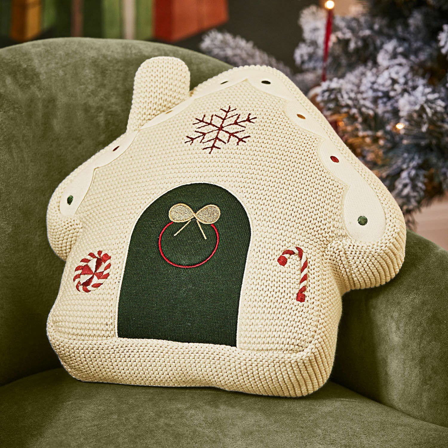 Stoff-Lebkuchenhaus Skavari Dekoratives Kissen in Hausform aus cremefarbenem Strick mit Weihnachtsmotiven und grünen Details auf grünem Sessel