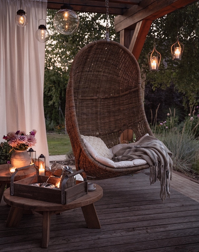 Hängesessel aus braunem Rattan auf Terrasse mit Kissen und Decke, beleuchtet von dekorativen Lampen