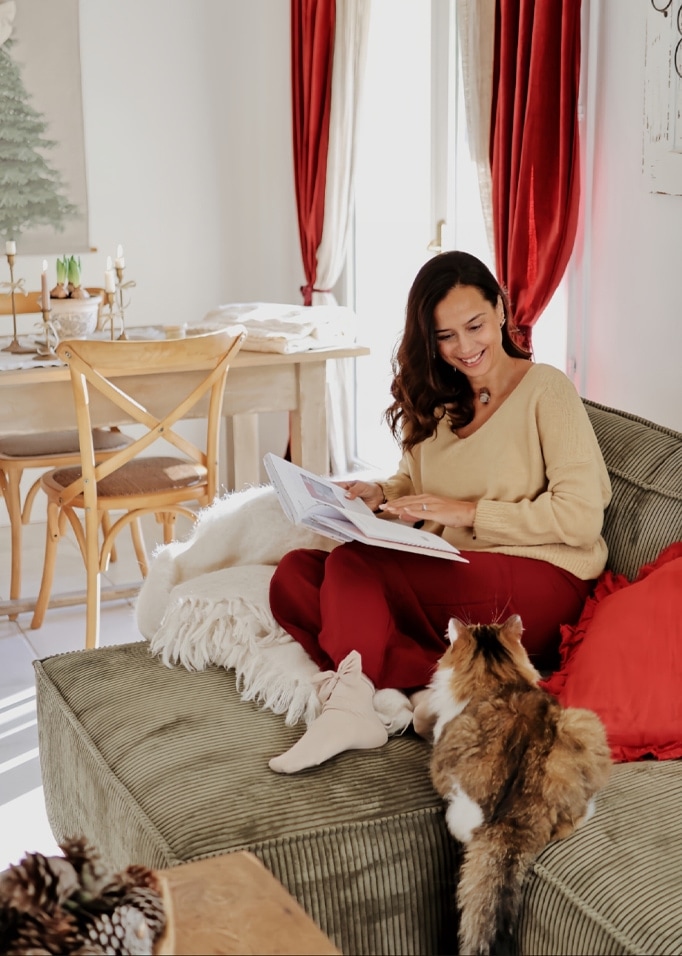 Frau in beigem Pullover und roter Hose liest Buch auf grünem Sofa mit Katze in gemütlichem Wohnzimmer