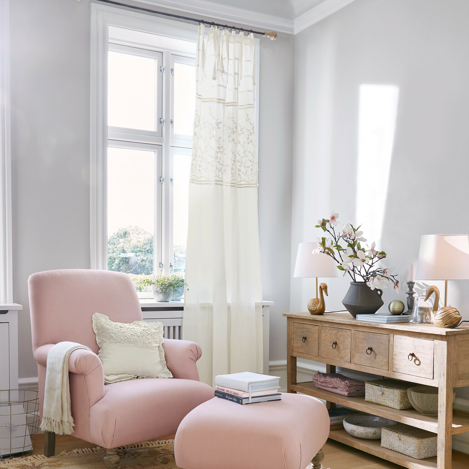 Rosa Sessel mit Hocker und cremefarbenem Kissen in hellem Wohnzimmer mit Holzkommode und Dekorationen