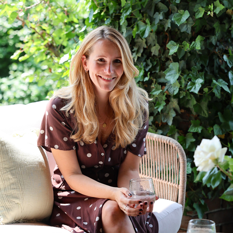 Frau in braunem gepunktetem Kleid sitzt mit Glas in der Hand auf einem Korbsessel im Garten