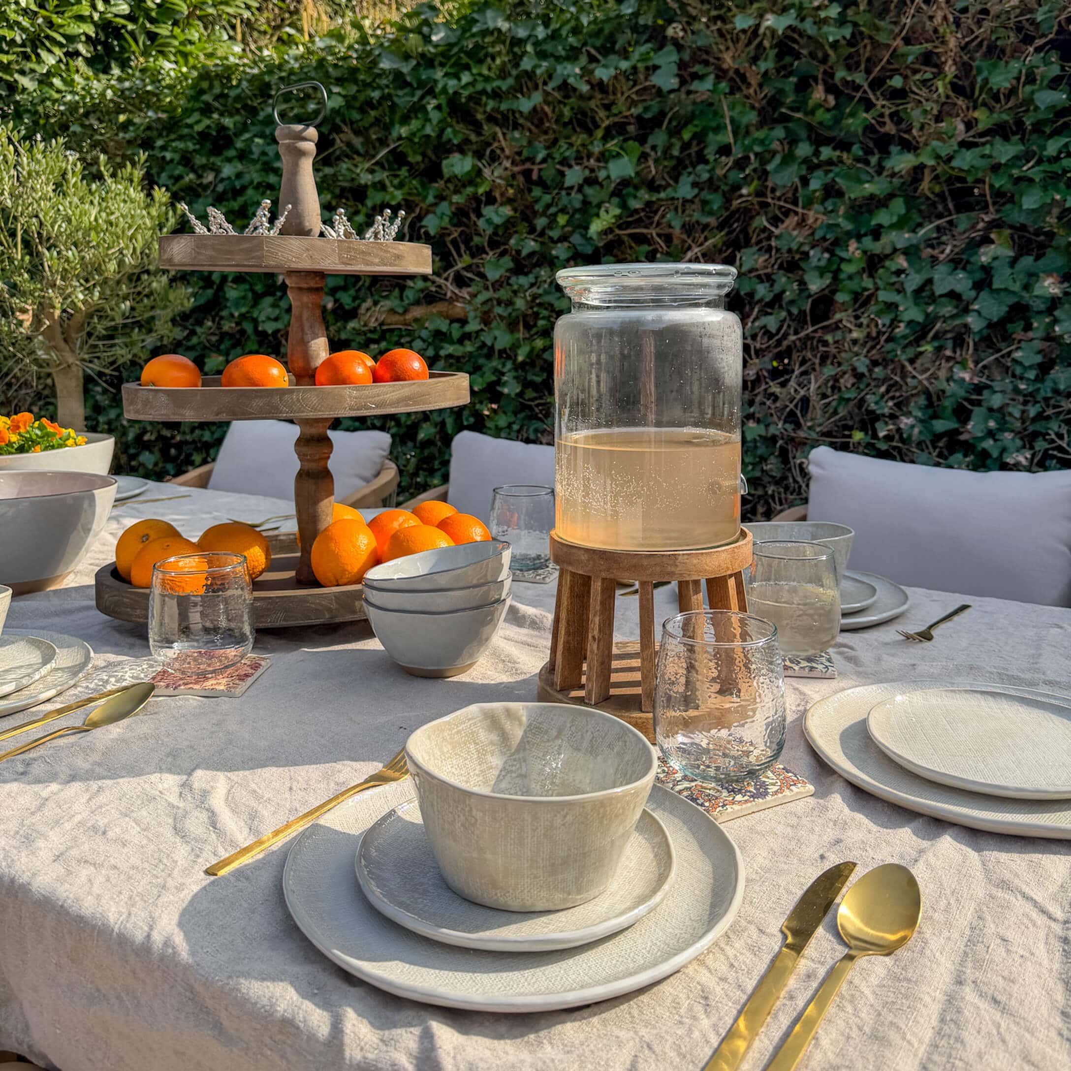 Gartentisch mit cremefarbener Keramik, goldfarbenem Besteck und hölzernem Obstständer