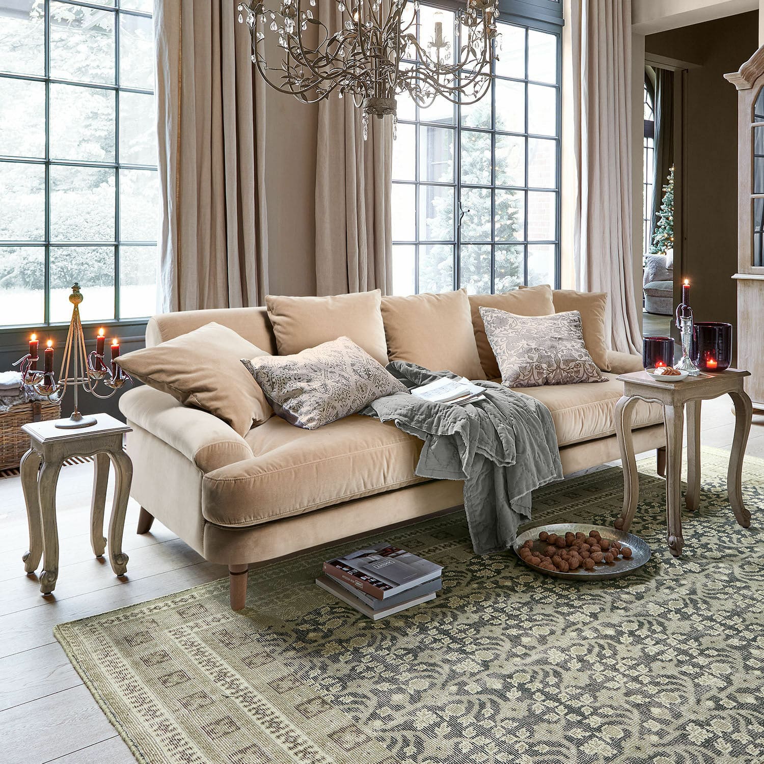 Sofa Rivermere Beige Samtsofa mit Kissen und grauer Decke im hellen Wohnzimmer mit Teppich und Kerzen