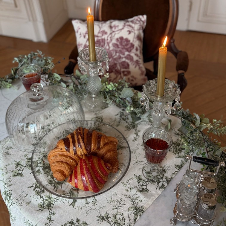 Glasteller mit Croissant und Gebäck auf Tisch mit weißem Tischtuch, zwei goldene Kerzen in Kristallkerzenhaltern