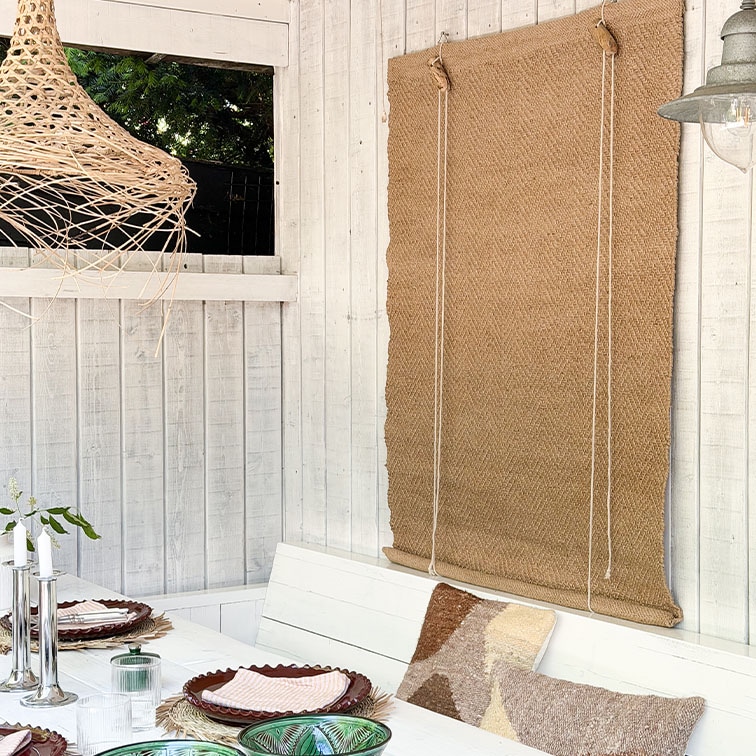 Weiße Holzwand mit natürlichem Jute-Rollo und dekorativem Tisch mit Kissen im Boho-Stil