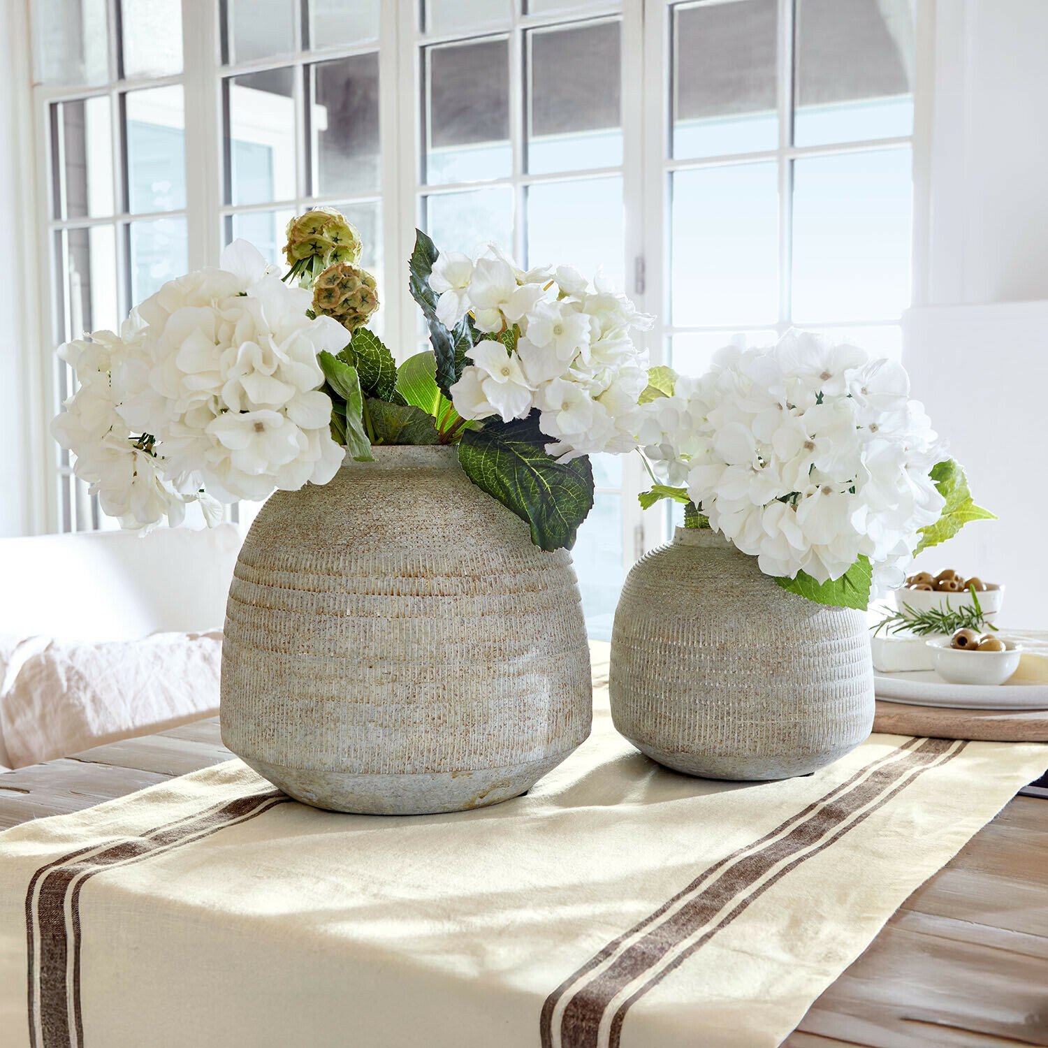 Zwei graue Keramikvasen mit weißen Blumen auf beigem Tischläufer in hellem Raum
