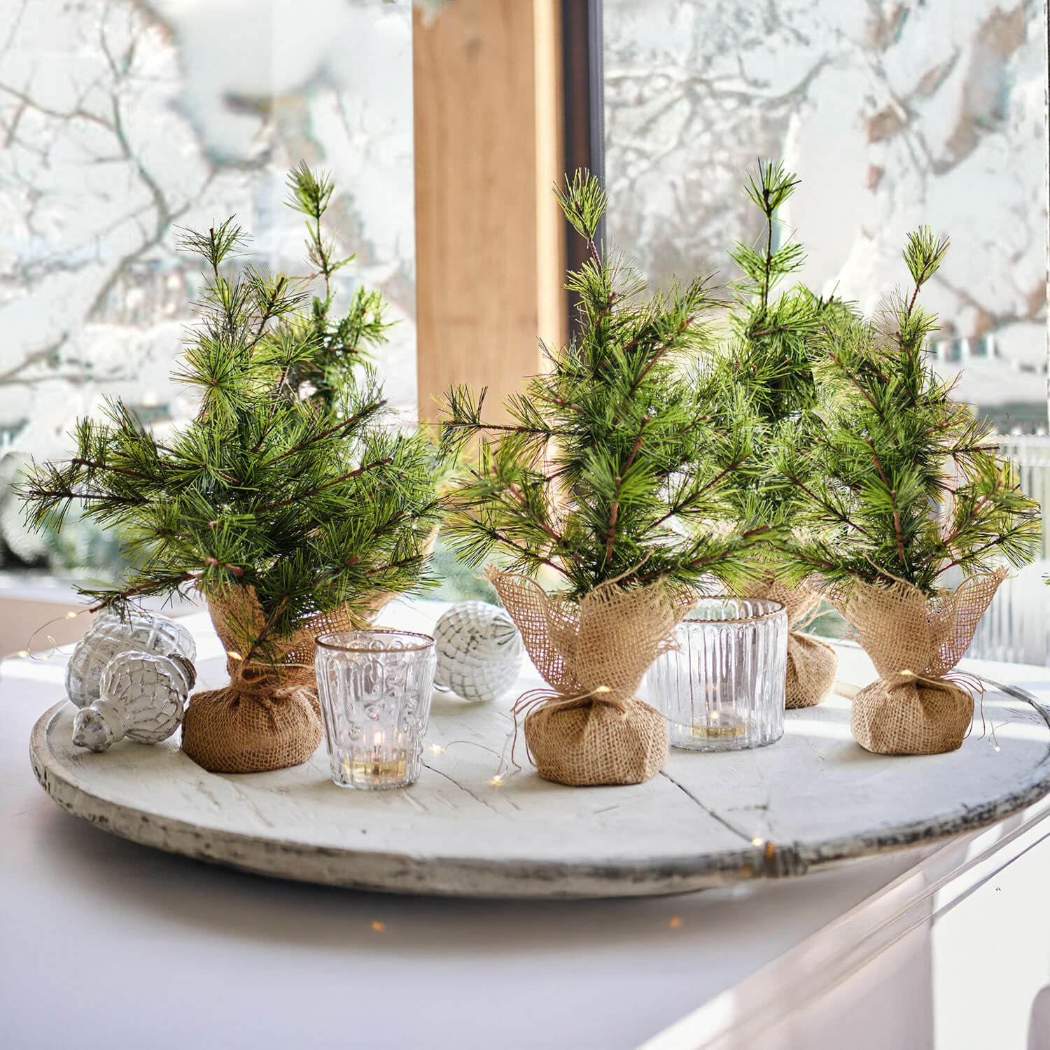 Kleine dekorative Tannenbäume in Jutesäcken auf weißem Tablett mit Kerzen und Weihnachtsdeko