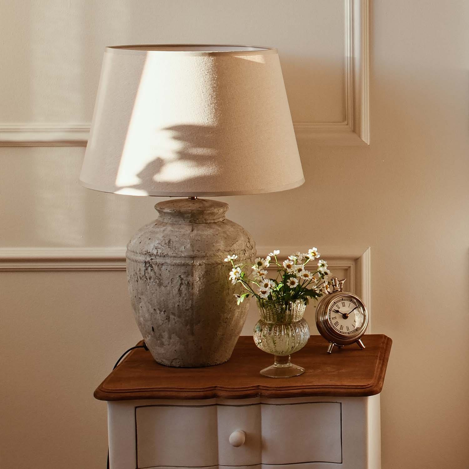Tischlampe mit beigem Stoffschirm und vintage Keramikfuß auf Holztisch mit Blumen und Wecker