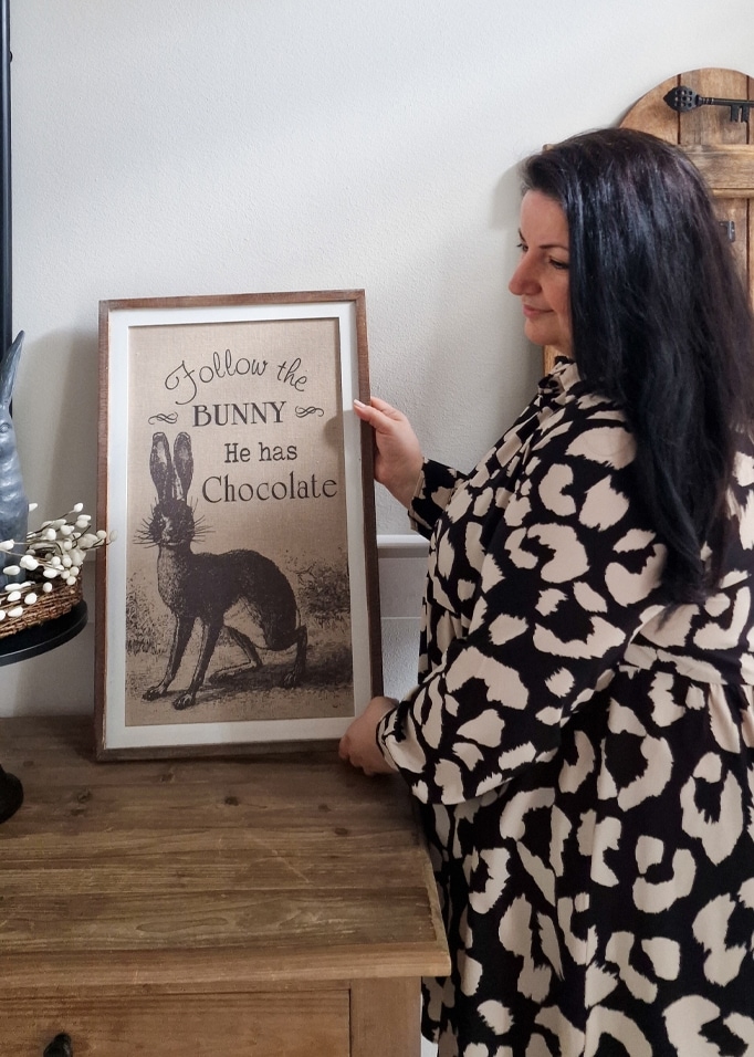 Frau hält gerahmtes Bild mit Hasenmotiv und Schokoladentext auf einem Holztisch