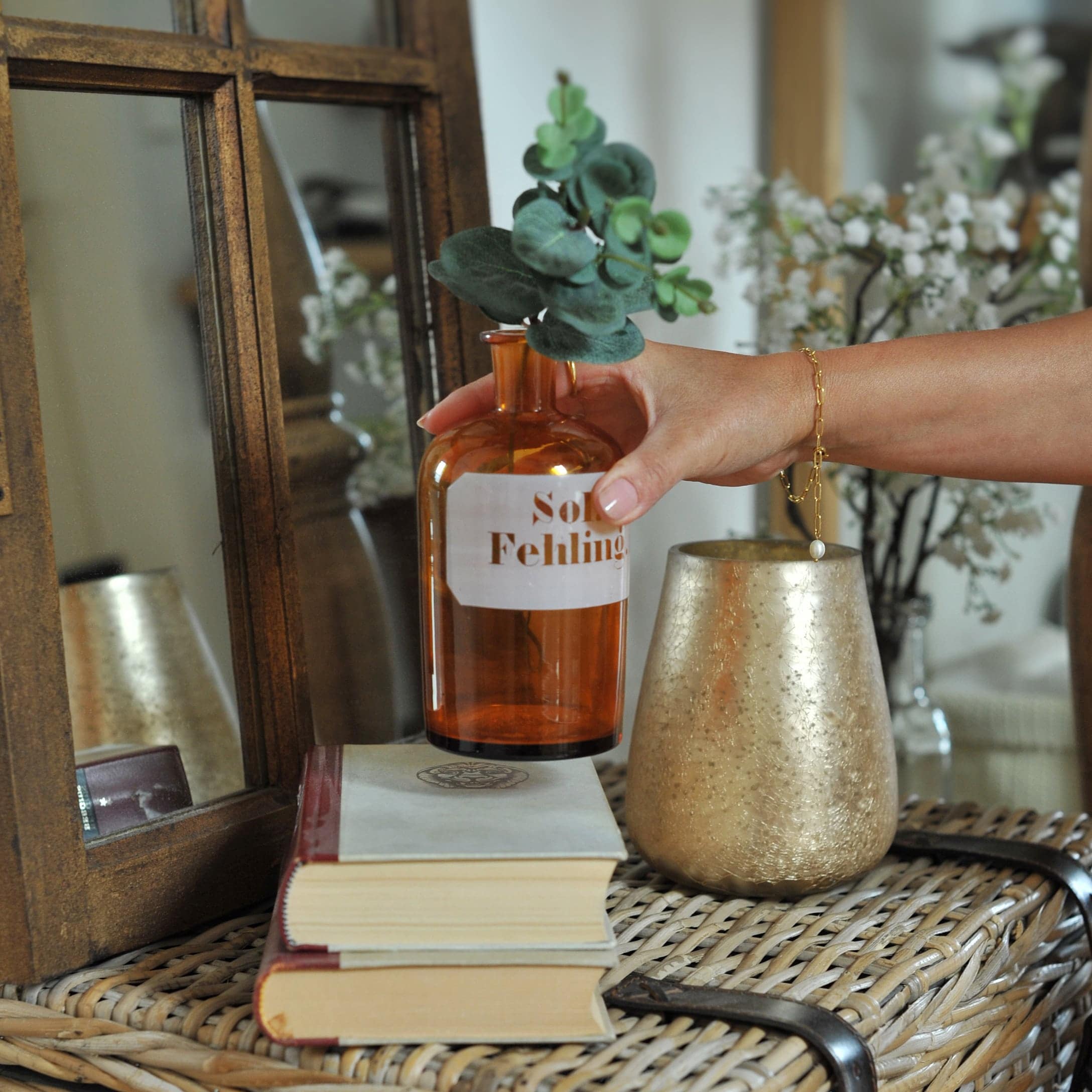 Hand hält bernsteinfarbene Glasflasche mit grünen Blättern auf Rattantisch mit Büchern und goldenem Kerzenhalter