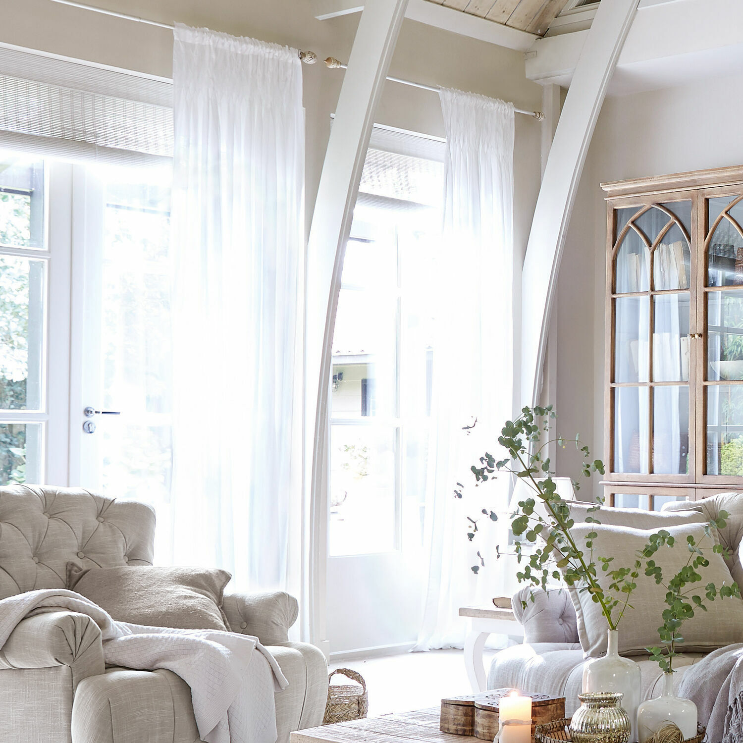 Gardine Otilie Helles Wohnzimmer mit beigem Polstersessel, weißen Vorhängen und vintage Holzvitrine