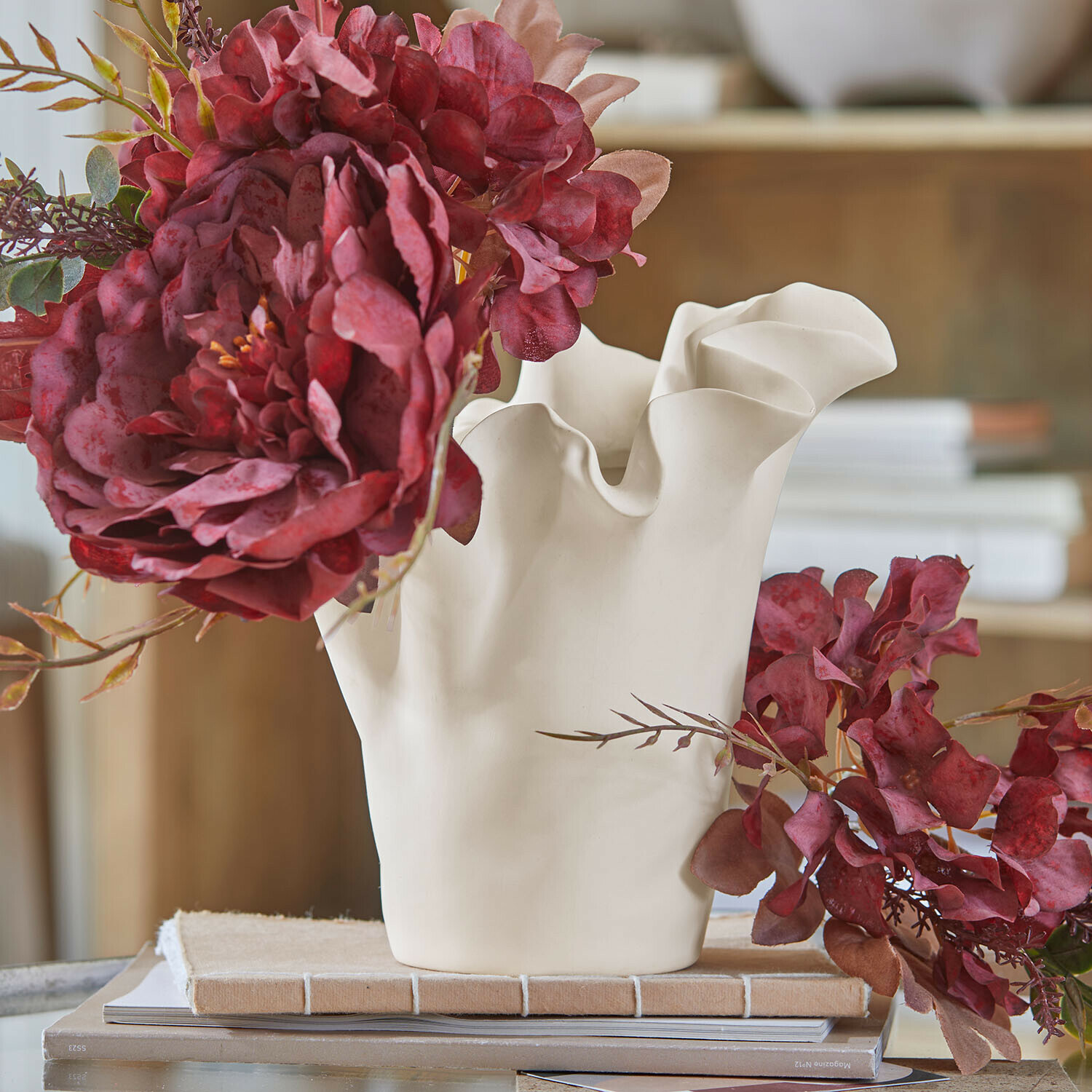 Weiße Keramikvase mit wellenförmigem Rand und roten künstlichen Blumen auf einem Tisch mit Büchern