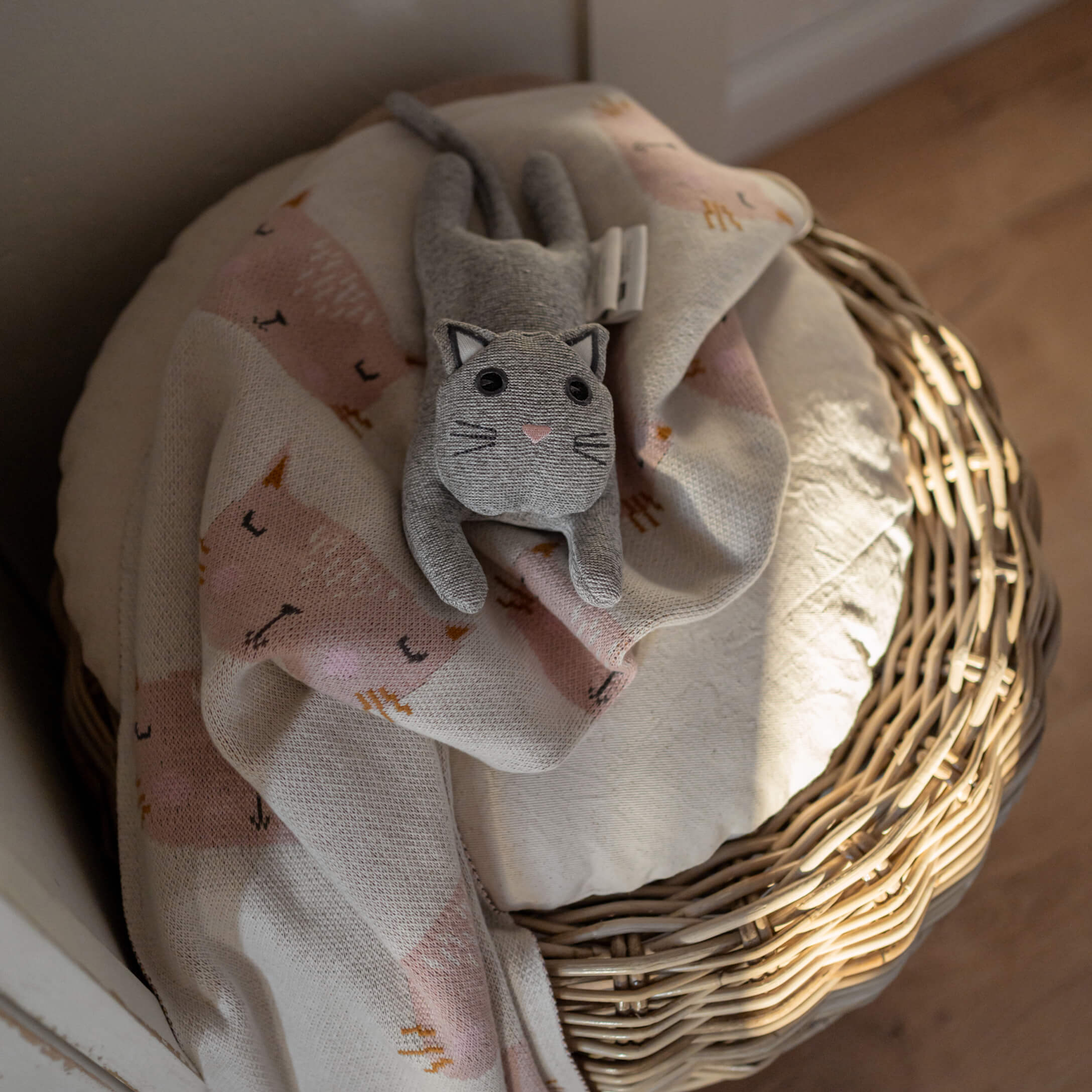 Graue Plüschkatze auf hellrosa Decke mit Katzenmotiv auf einem Korb aus Weide