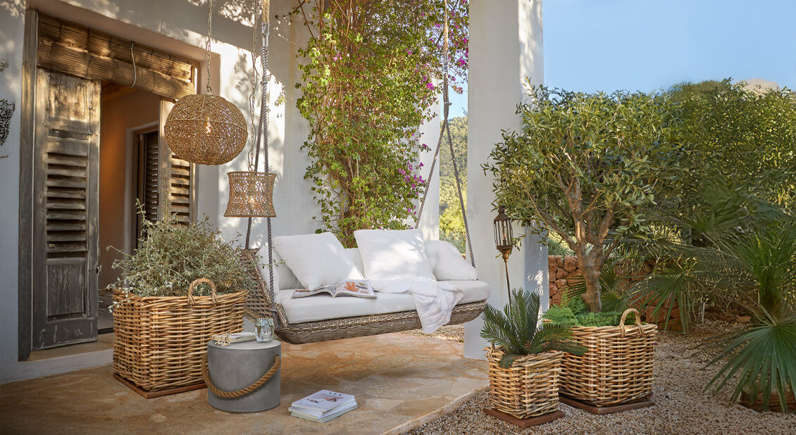 Wikling-Schaukel mit weißen Kissen auf Terrasse mit Rattanlampen und Pflanzen in geflochtenen Körben