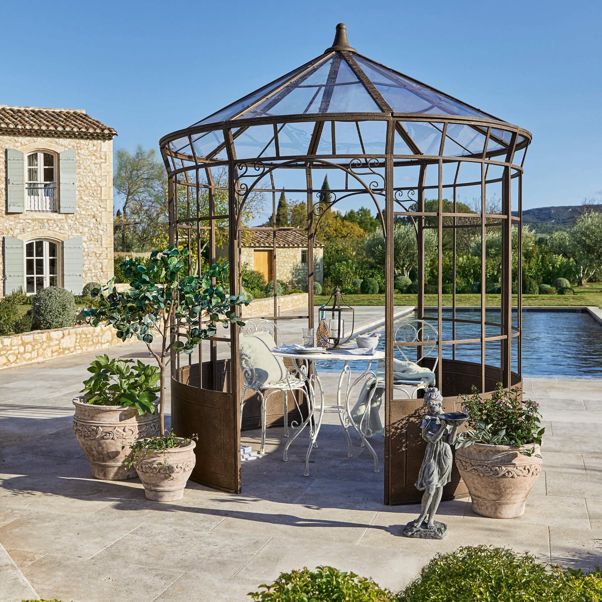 Garten mit rostfarbener Metalllaube, weißem Tisch und Stühlen, umgeben von Pflanzgefäßen