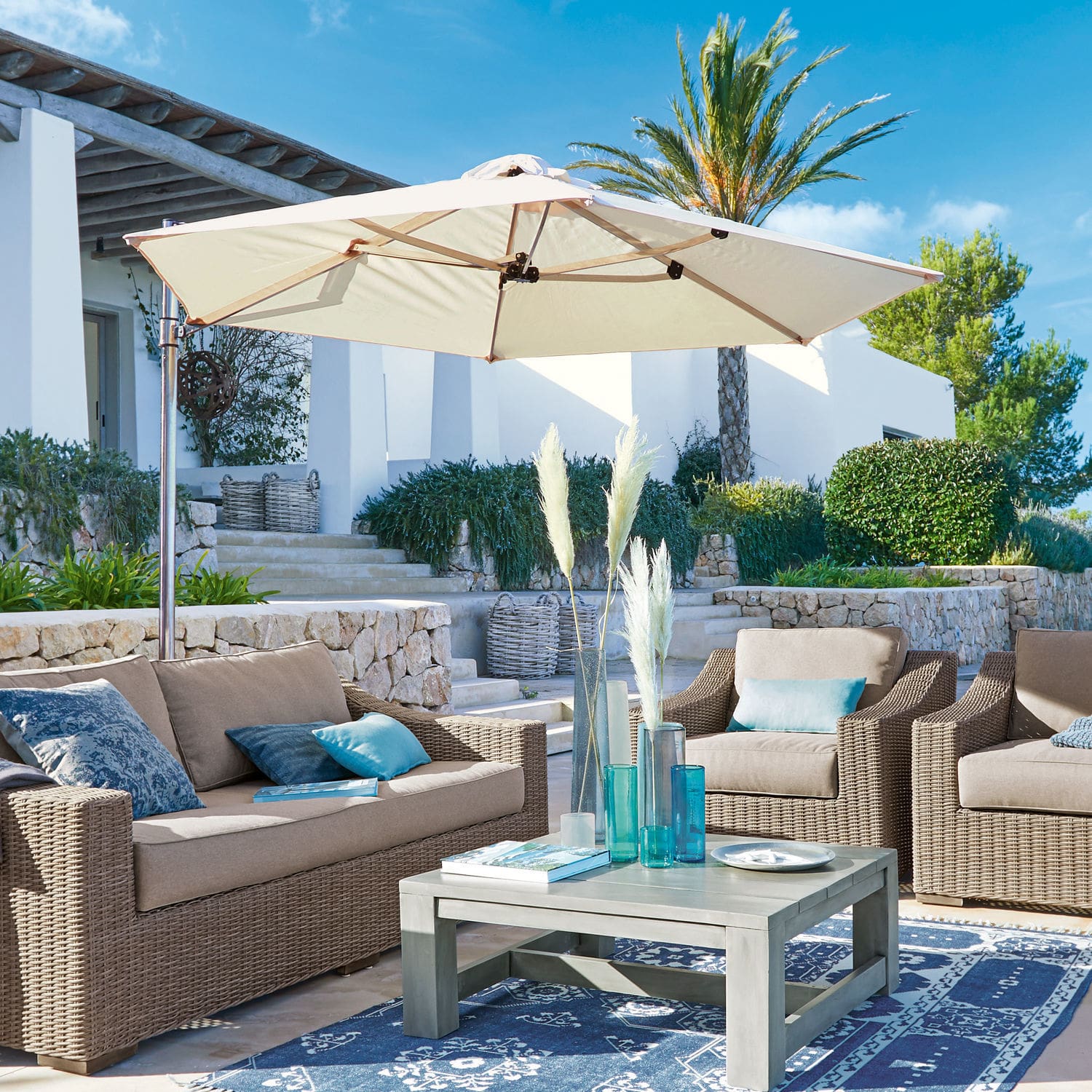 Sonnenschirm Boudicca Gartenmöbel-Set aus beigem Polyrattan mit cremefarbenem Sonnenschirm auf Terrasse mit blauem Teppich