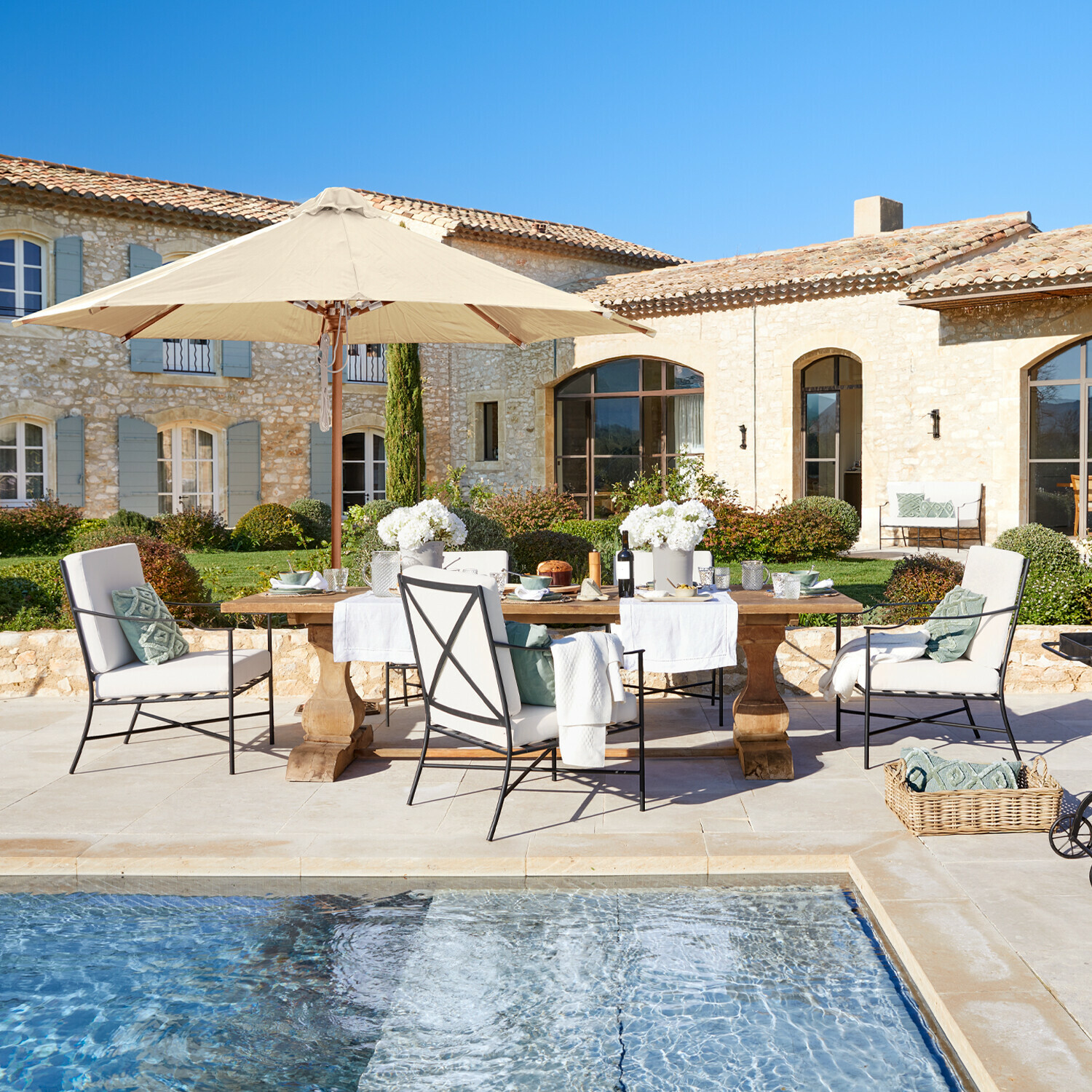 Tisch Devonshire Gartenmöbel-Set mit beigem Sonnenschirm und Kissen am Pool vor einem Steinhaus