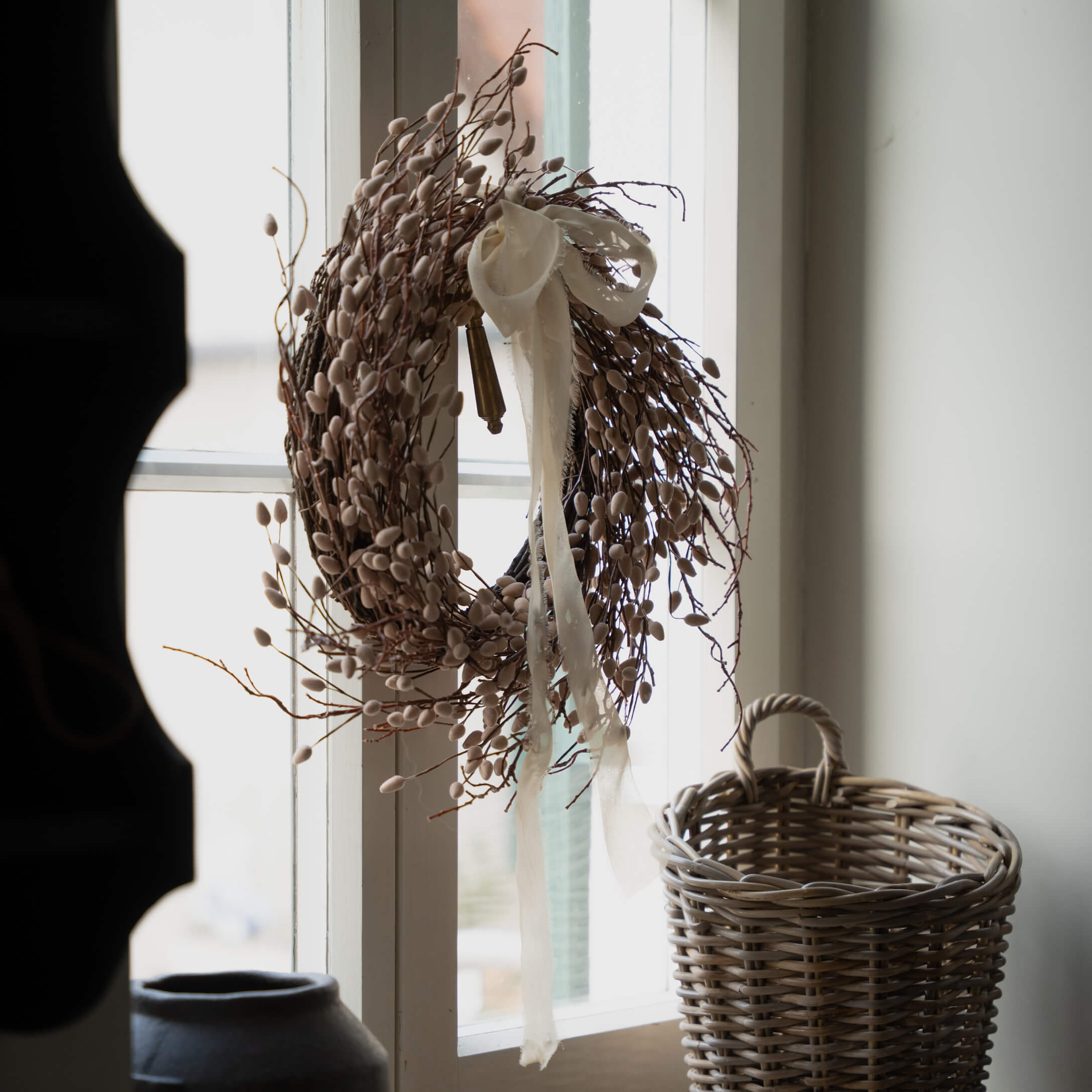 Dekorativer Kranz aus Zweigen und Knospen in Beige mit weißem Band am Fenster, daneben ein Korb aus Weide