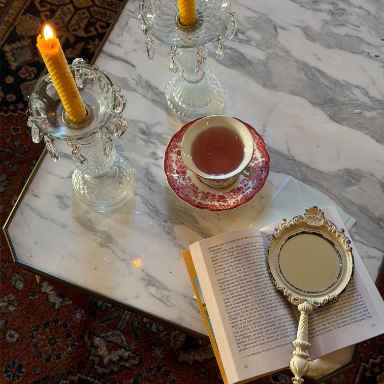 Gläserne Kerzenhalter mit gelben Kerzen und Tasse mit rotem Muster auf Marmorplatte mit Buch und Handspiegel