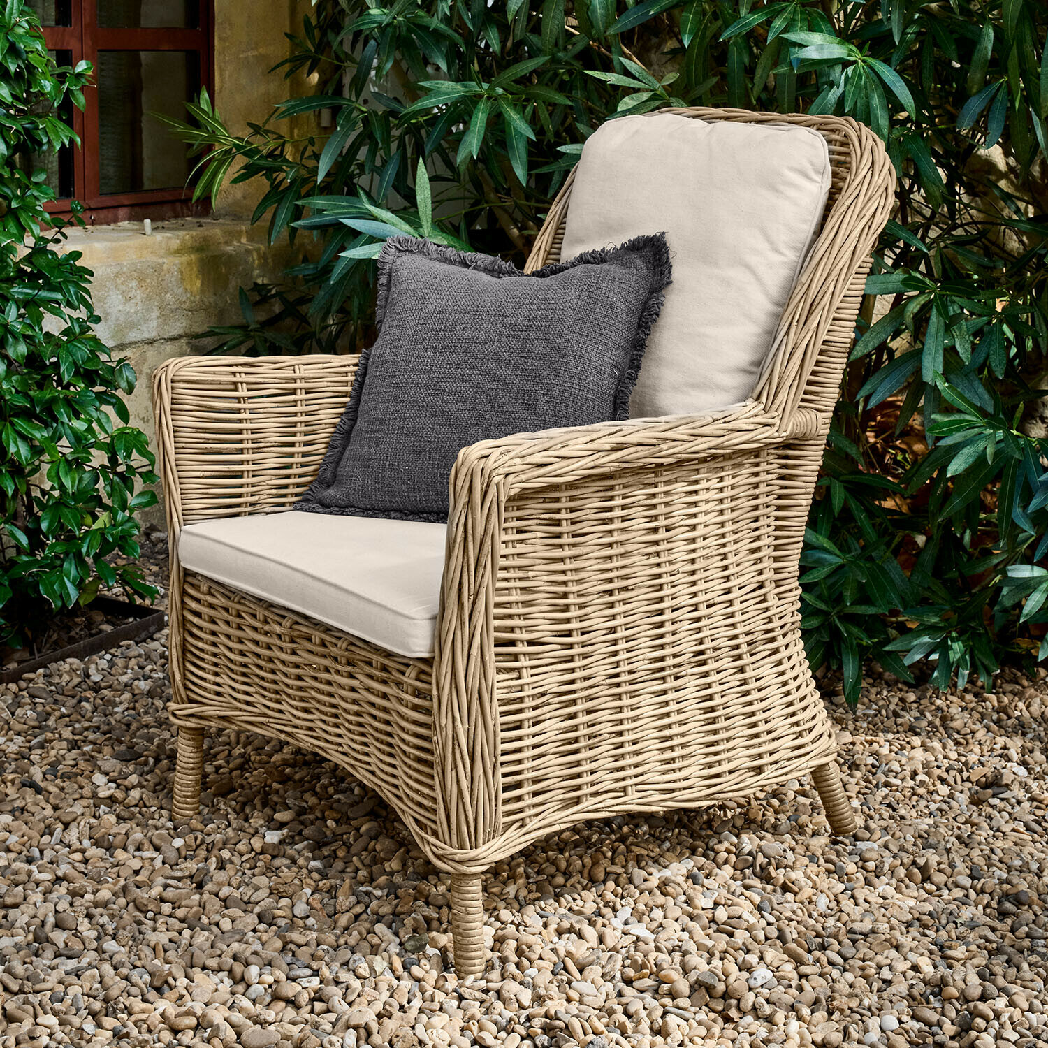 Sessel Travenne Rattansessel mit cremefarbenen Polstern und dunkelgrauem Dekokissen auf Kieselsteinboden im Garten