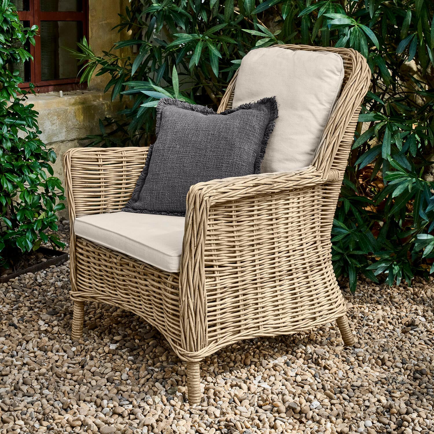 Sessel Travenne Rattansessel mit cremefarbenen Polstern und dunkelgrauem Dekokissen auf Kieselsteinboden im Garten