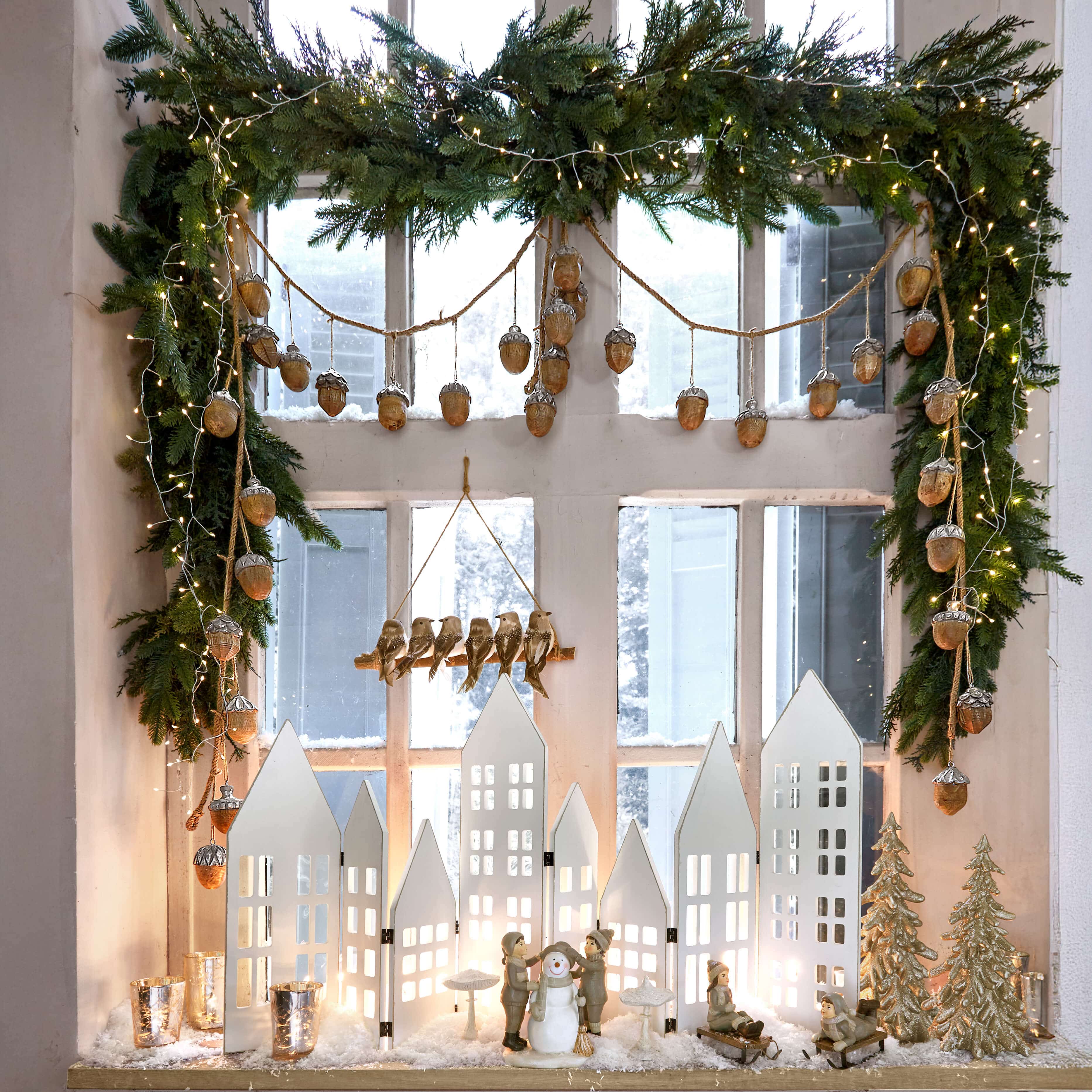 Weihnachtsdekoration mit weißen Häusern, Kinderfiguren und Schneemann auf Fensterbank mit grüner Girlande und Lichterkette