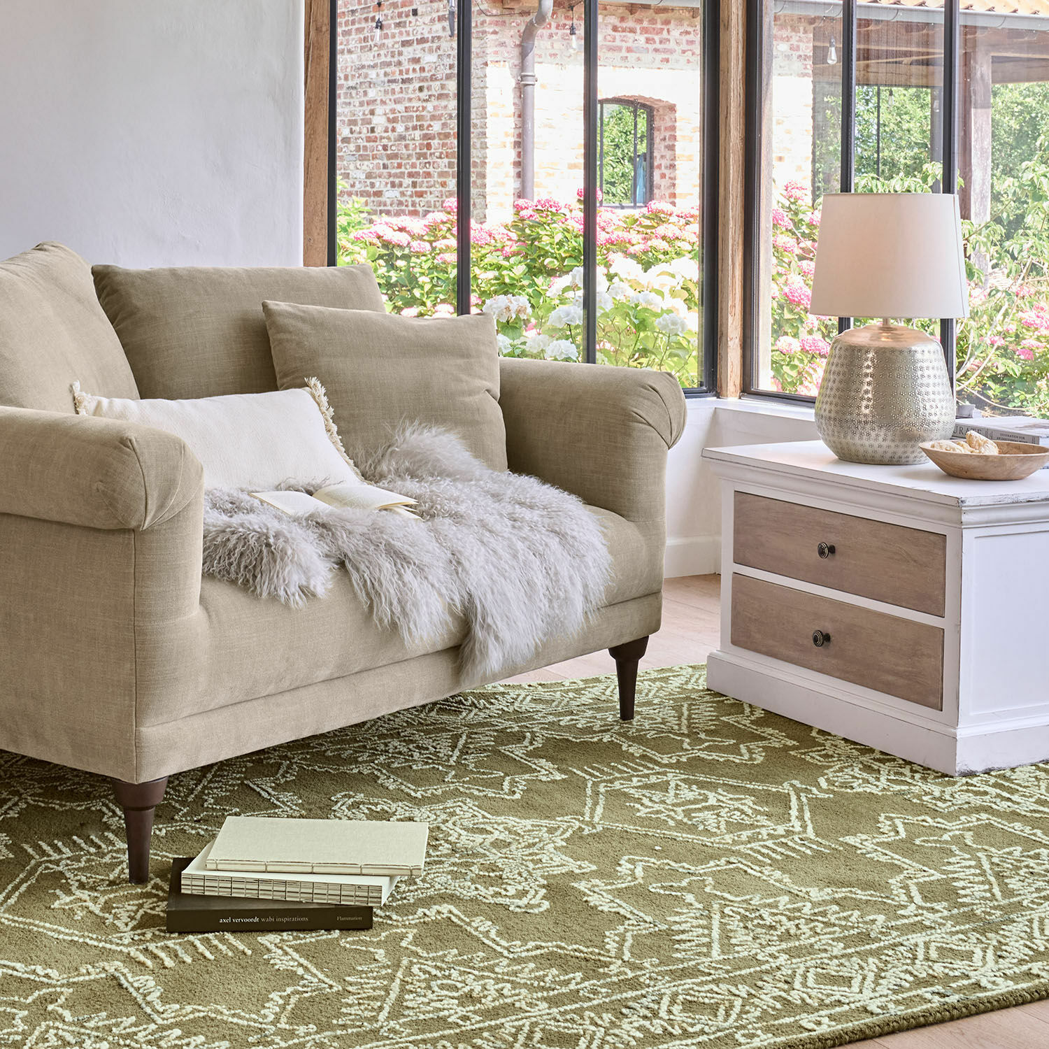 Teppich Qazoura Beiges Stoffsofa mit Kissen und Fellüberwurf auf grünem Teppich mit geometrischem Muster