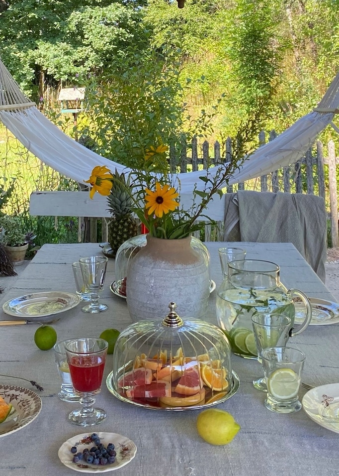 Gartentisch mit grauem Tuch, Vase mit gelben Blumen und Glasglocke mit frischen Früchten.