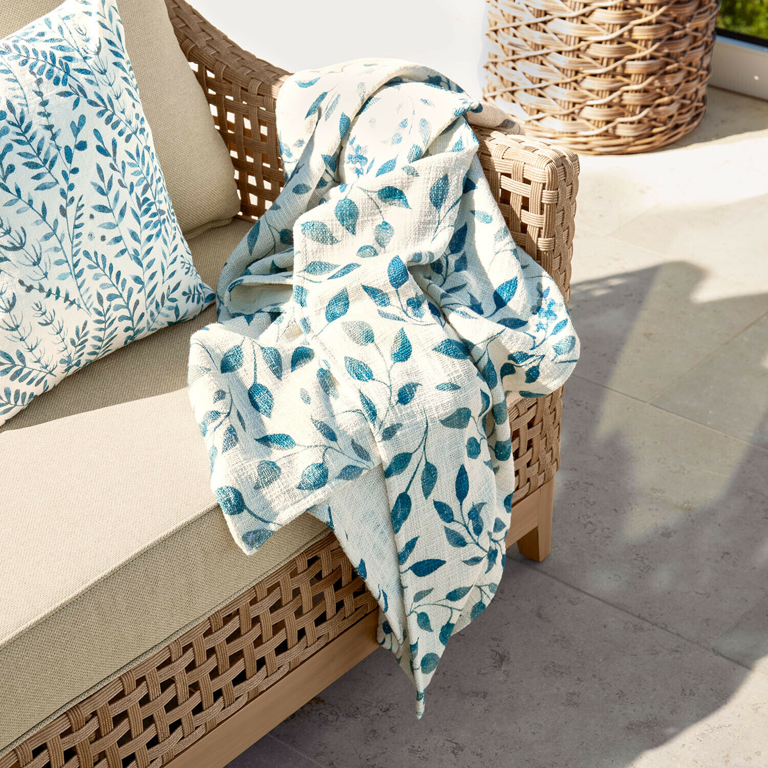 Beiges Sofa mit geflochtenem Rücken, darauf cremefarbene Decke mit blauem Blattmuster