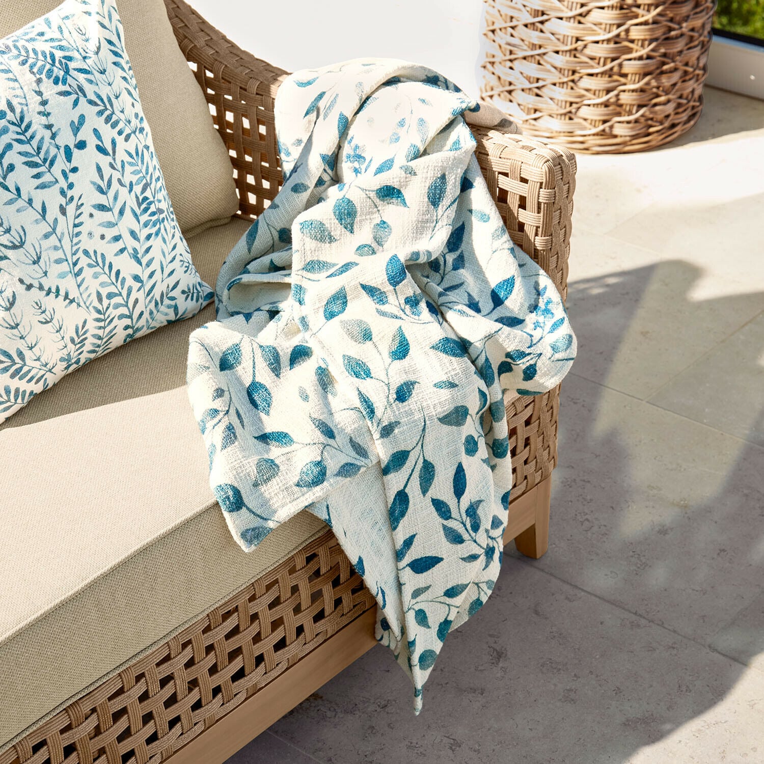 Plaid Ocevara Beiges Sofa mit geflochtenem Rücken, darauf cremefarbene Decke mit blauem Blattmuster